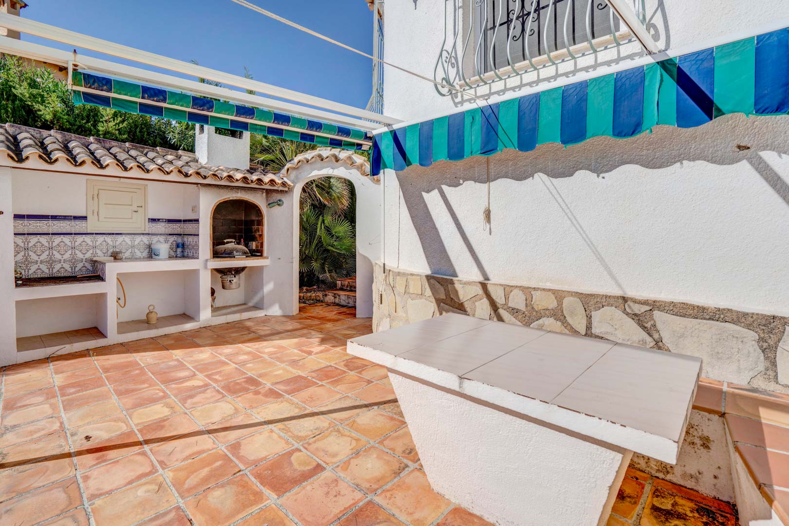 Zona exterior con barbacoa de obra decorada con azulejos tradicionales, mesa fija, toldos extensibles y suelos de terracota en un entorno mediterráneo.