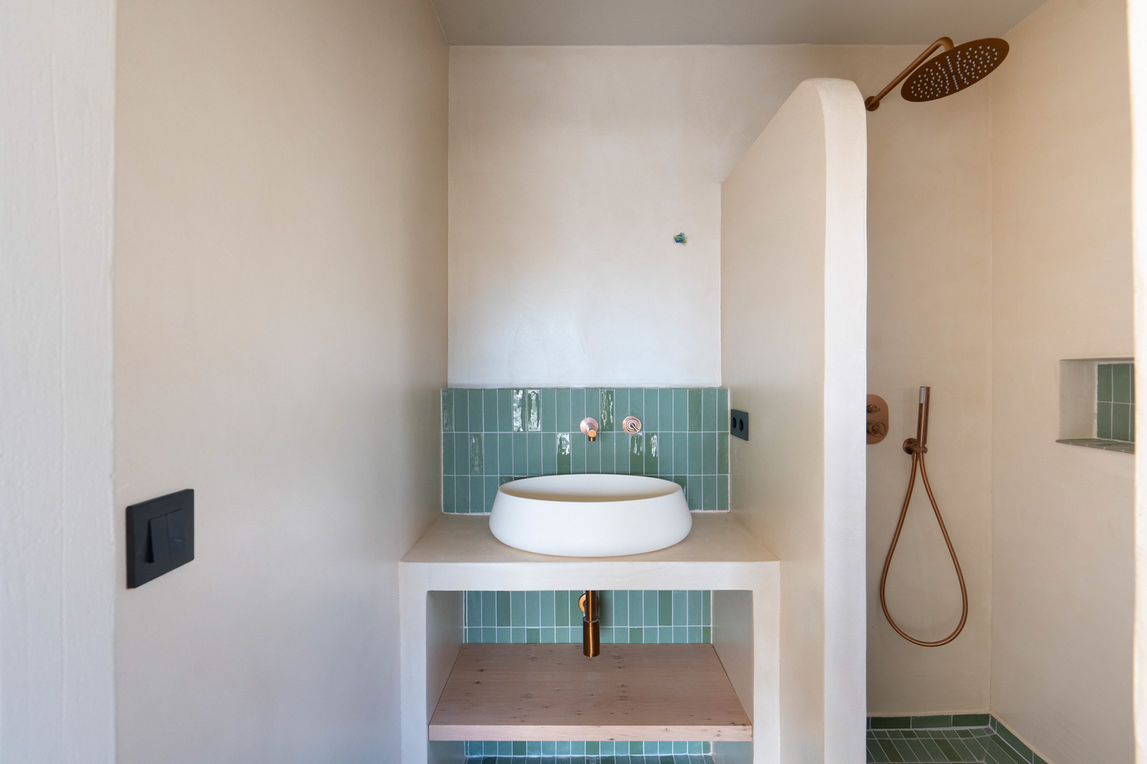 Baño con lavabo sobre encimera, grifería de cobre y azulejos verdes verticales. Paredes de arcilla y ducha de obra con una característica mampara curva.