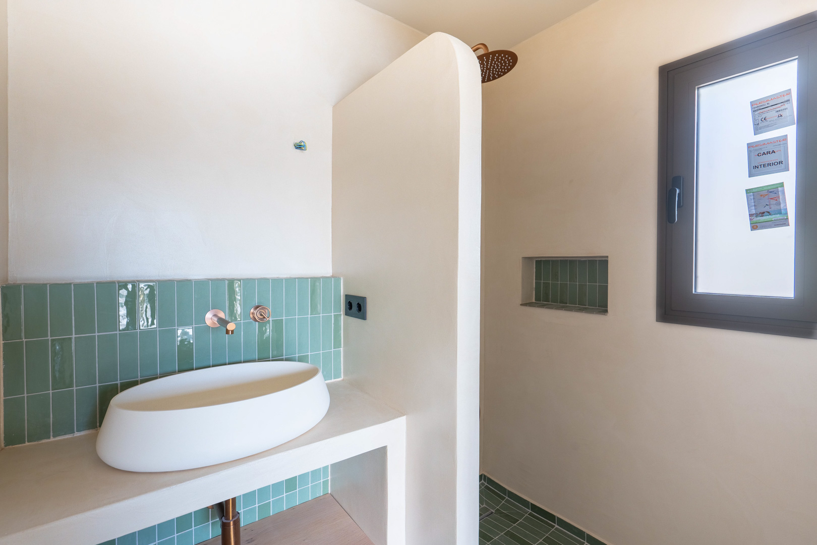 Baño de diseño con lavabo sobre encimera, grifería empotrada en cobre y revestimiento de azulejos verdes verticales. Ducha de obra con hornacina y acabados en arcilla natural.