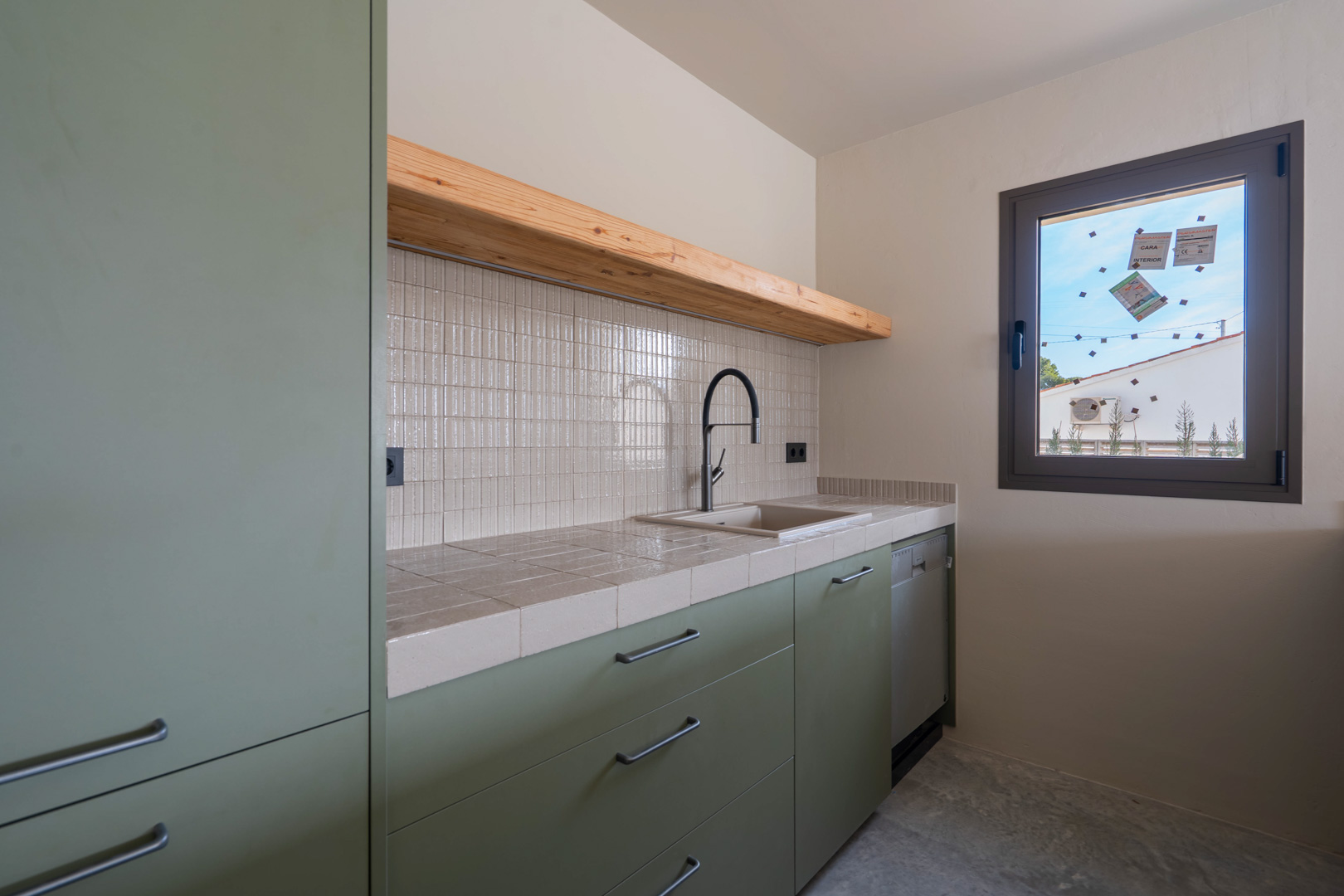 Cocina de invitados con armarios verde salvia, encimera de azulejos, estante de madera maciza y grifería negra sobre suelo de hormigón pulido.