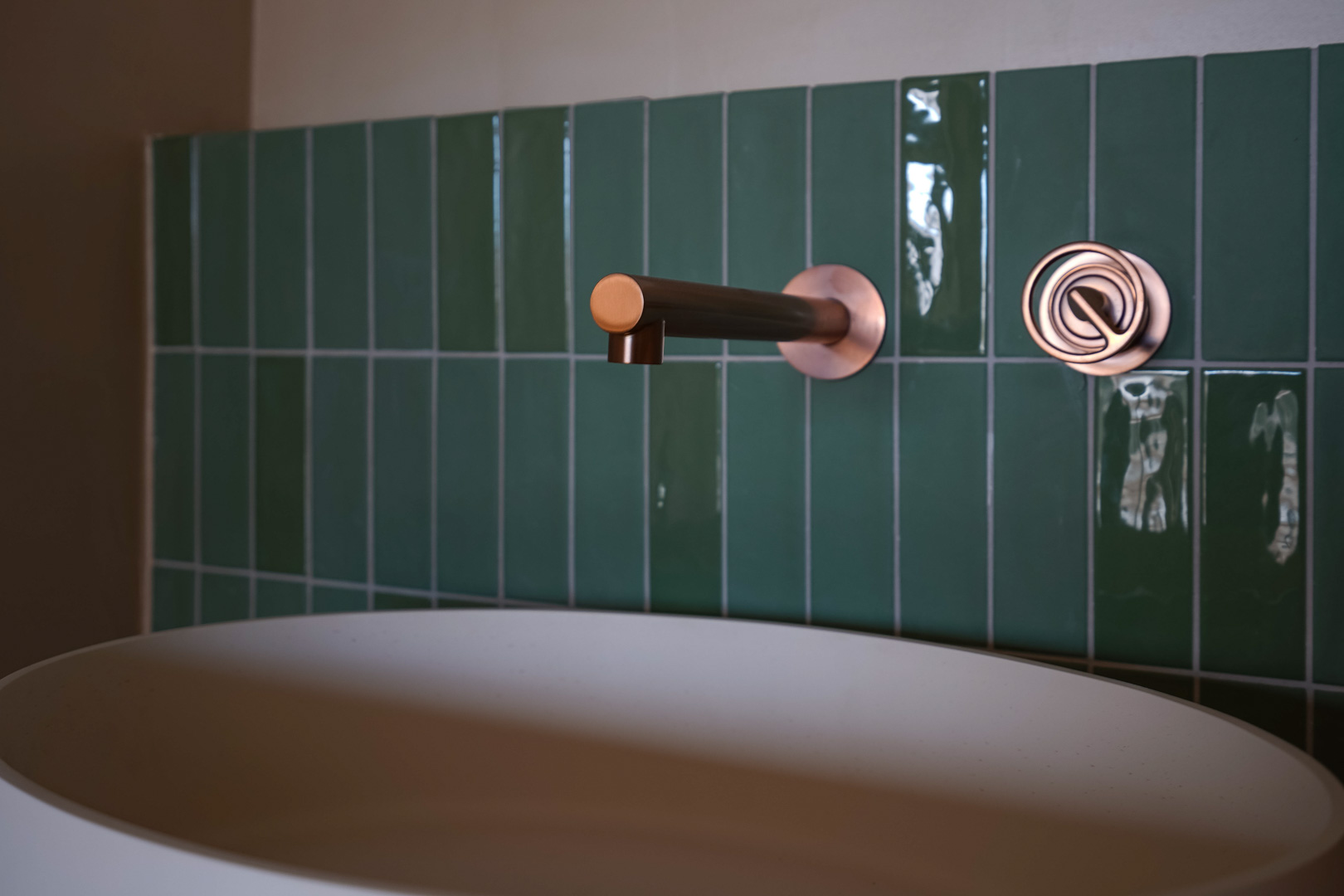Detalle de baño con grifería empotrada en acabado cobre sobre azulejos verticales de color verde esmeralda y lavabo de superficie blanco mate.