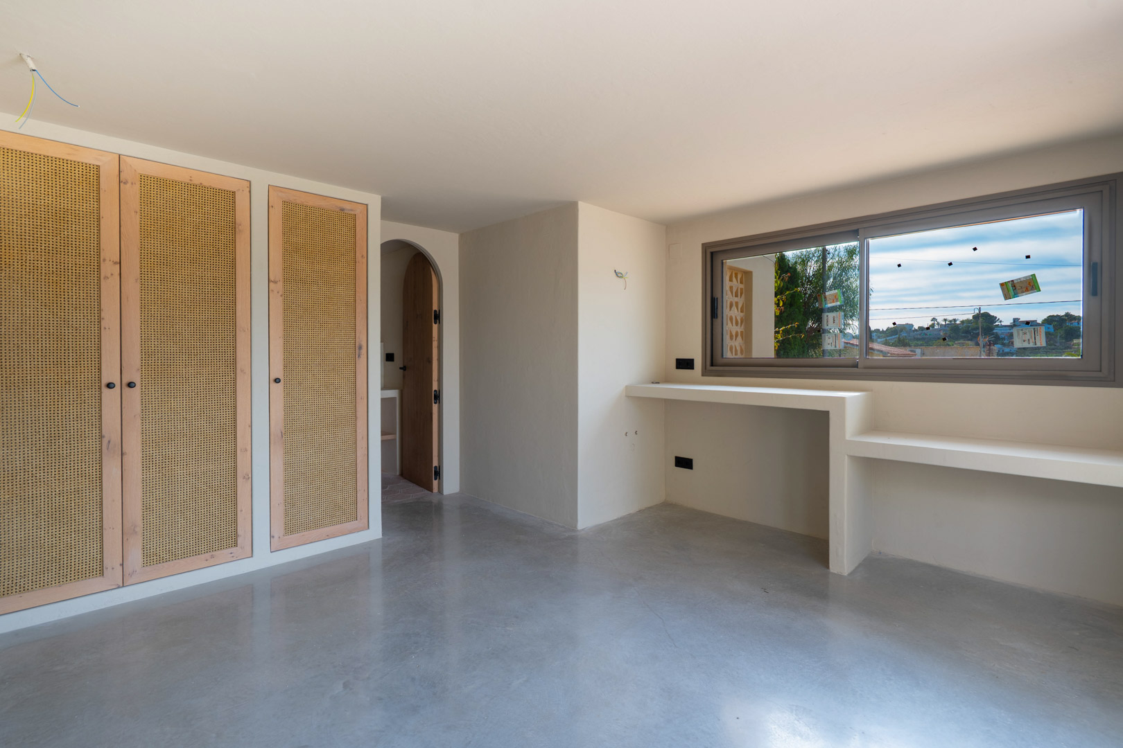 Dormitorio con armarios empotrados de madera y rejilla, suelo de hormigón pulido y zona de escritorio integrada bajo la ventana junto a un acceso en arco.