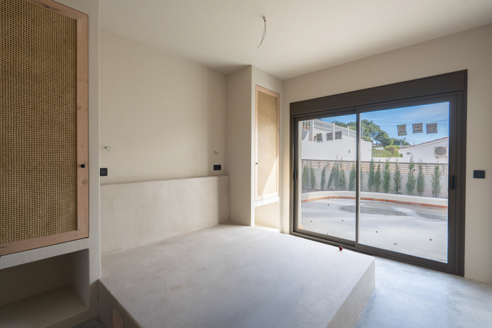 Dormitorio con base de cama en hormigón pulido, armarios empotrados con frontales de rejilla y acceso directo a la terraza mediante un gran ventanal corredero.