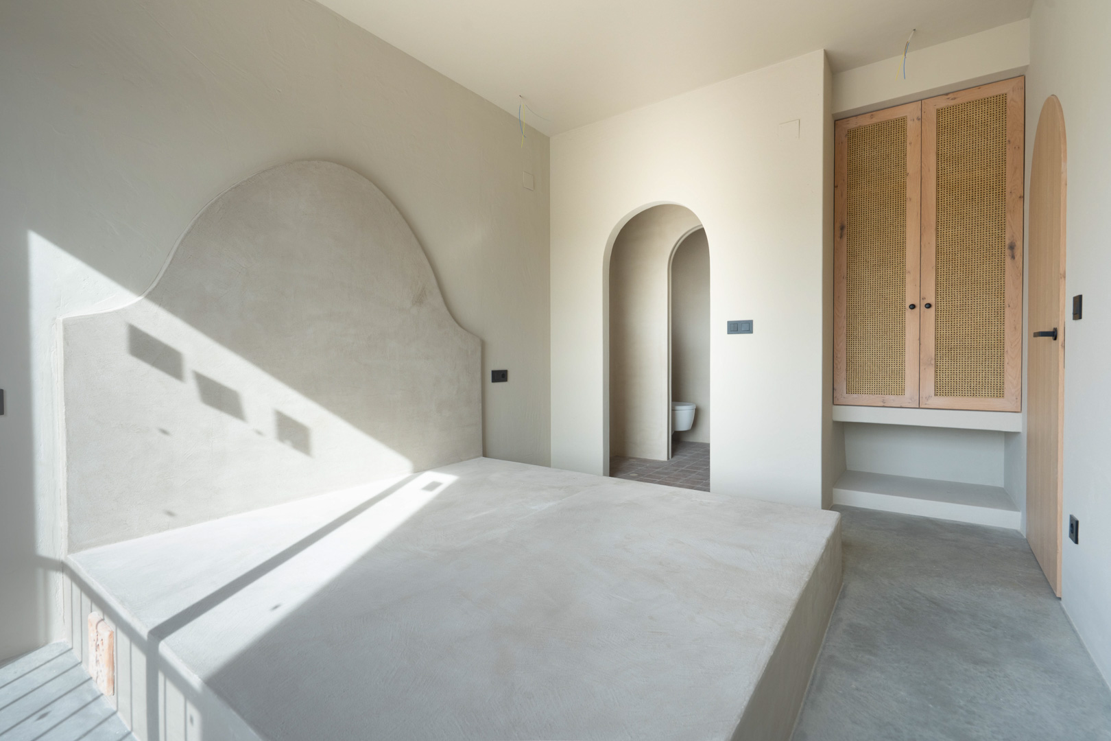 Dormitorio con base de cama integrada y cabecero de arcilla artesanal. Incluye armarios de roble con rejilla y acceso mediante arcos a un baño en suite.