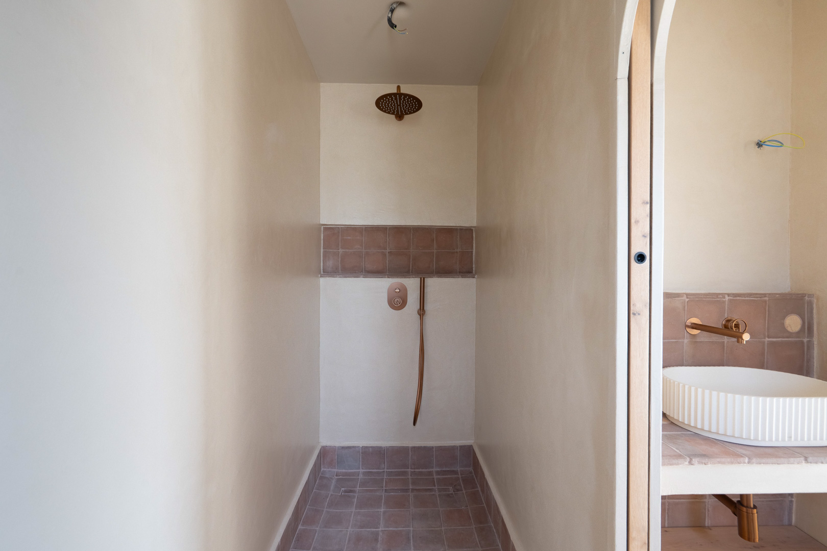 Ducha de obra con acabados en arcilla natural, grifería en tono bronce y suelo de terracota. Al fondo se aprecia el lavabo de diseño estriado sobre encimera.