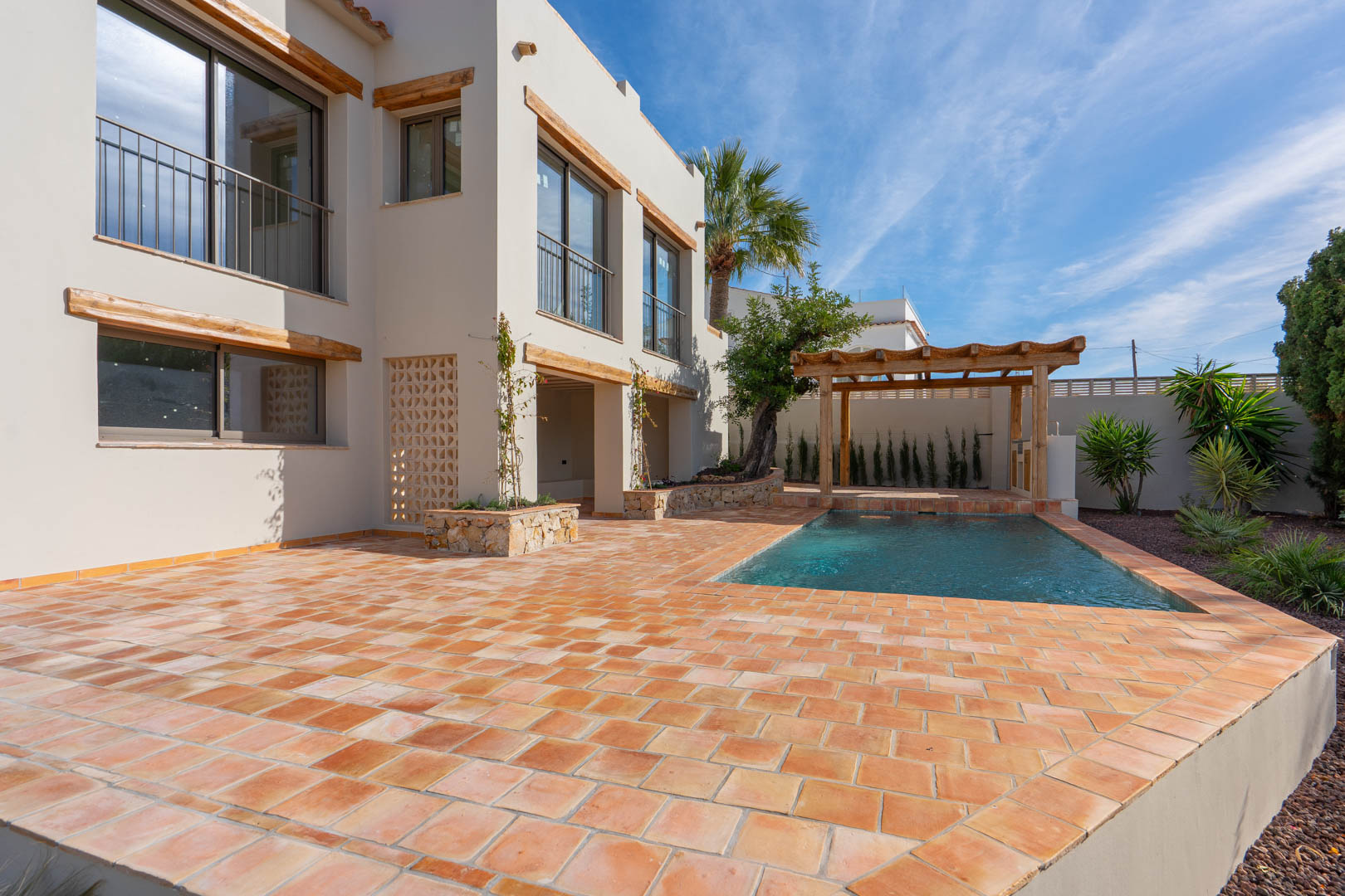 Exterior de la villa con amplia terraza de terracota, piscina de nueva construcción y pérgola de madera. La fachada destaca por sus dinteles de madera y acabados en piedra natural.