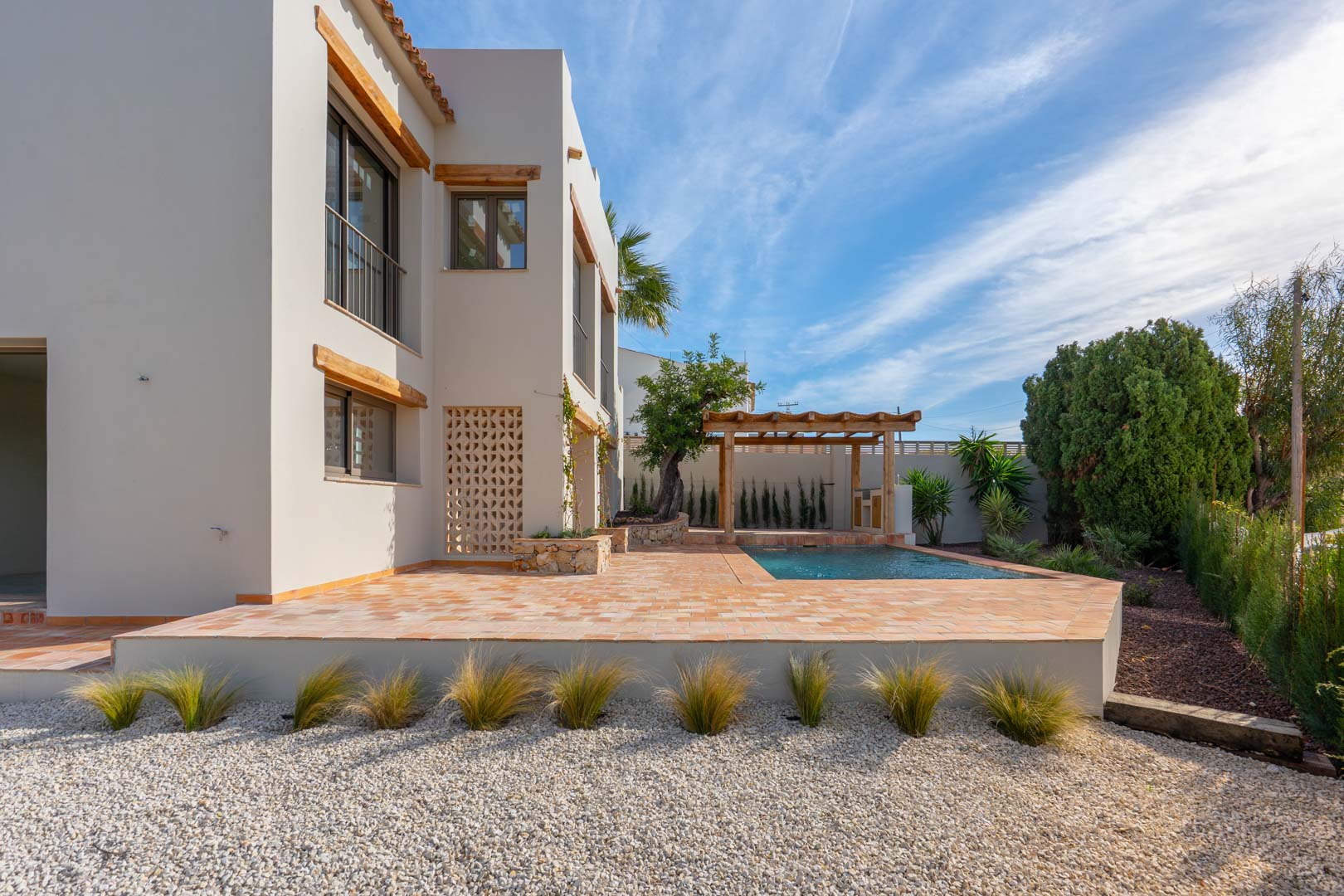 Exterior de la villa con terraza de terracota, piscina de nueva construcción y pérgola de madera, rodeada de un jardín mediterráneo con grava y vegetación ornamental.