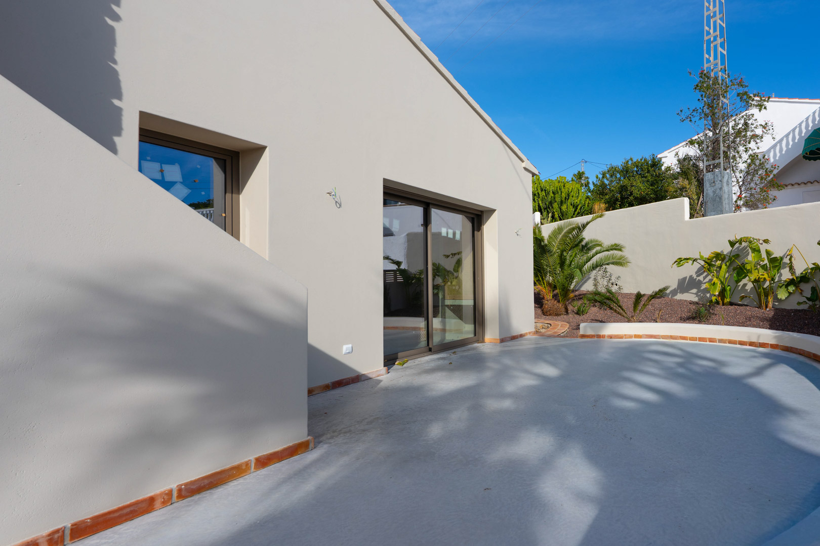 Fachada minimalista con acabados en tonos tierra, grandes ventanales y terraza curva con bordes de terracota junto a un jardín mediterráneo recién plantado.
