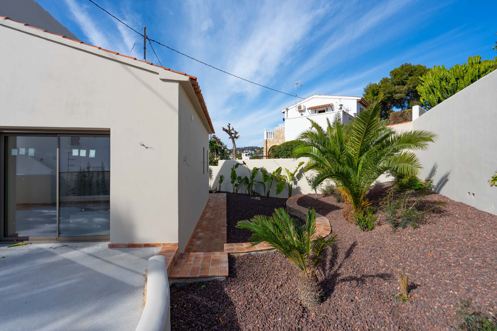Jardín mediterráneo con palmeras y grava volcánica, delimitado por senderos de terracota y muros blancos bajo un cielo despejado.