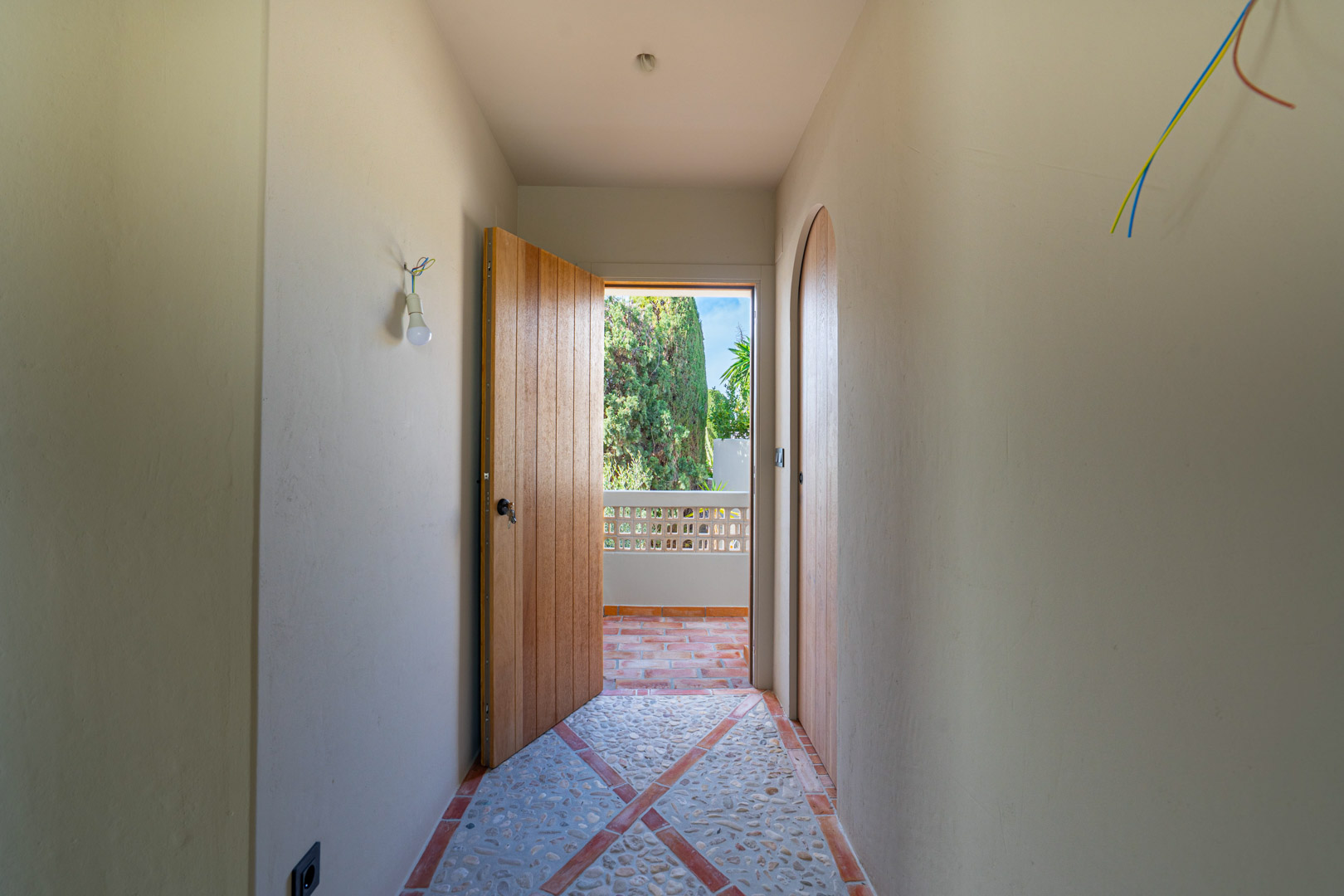 Pasillo con suelo de guijarros y terracota, paredes de arcilla y puertas de roble macizo que dan paso a una terraza con vistas al jardín mediterráneo.