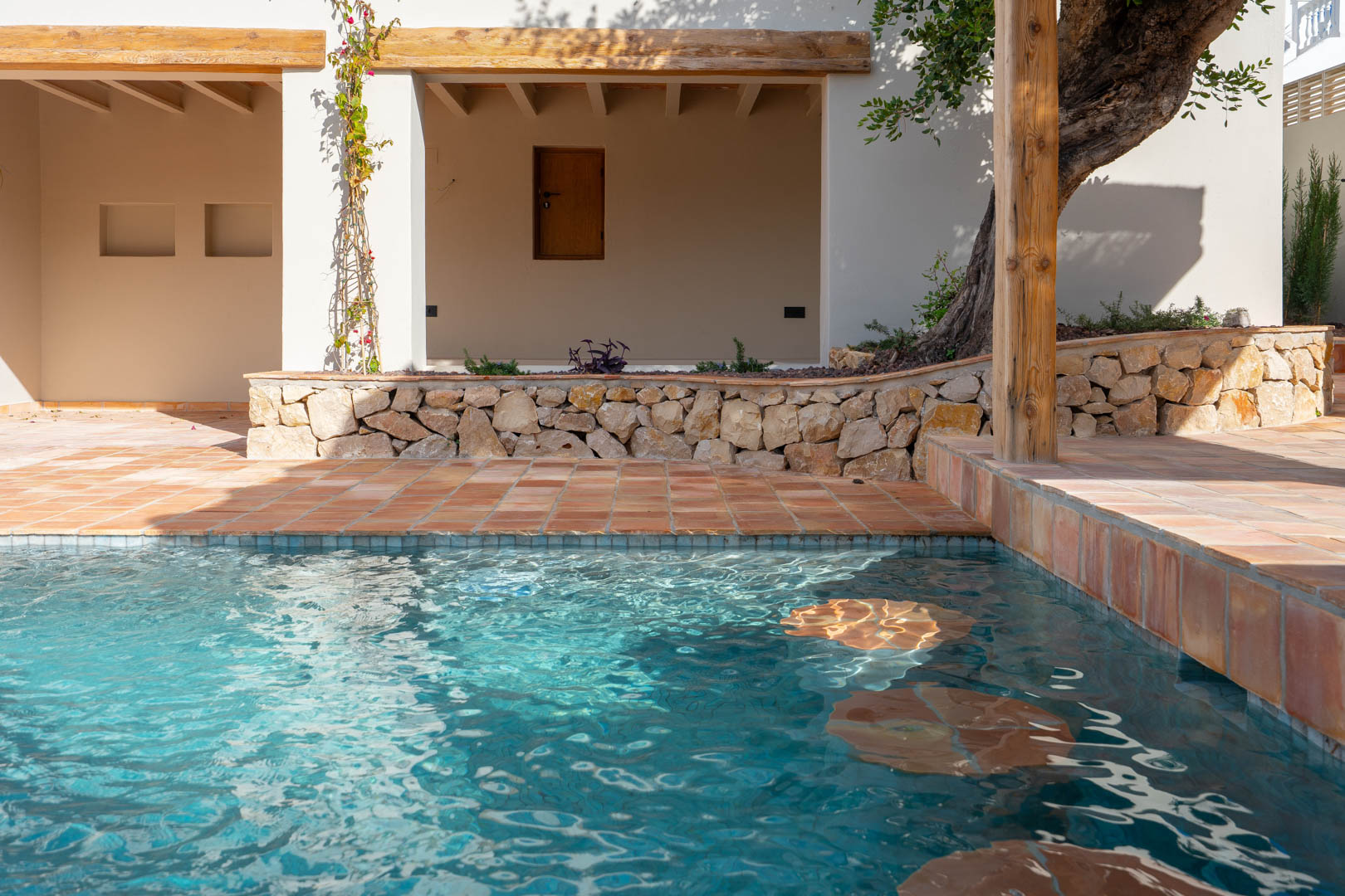 Piscina de nueva construcción con asientos integrados, rodeada de suelos de terracota, muros de piedra natural y vigas de madera maciza en la zona de porche.