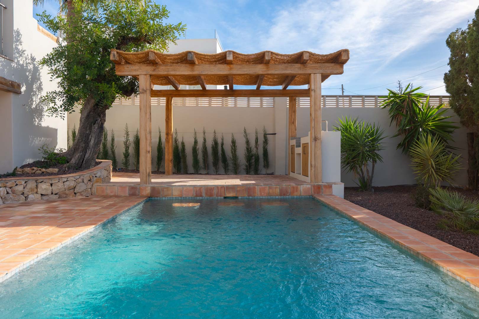 Piscina exterior con borde de terracota y una pérgola de madera rústica con zona de barbacoa integrada, rodeada de vegetación mediterránea y muros de piedra natural.
