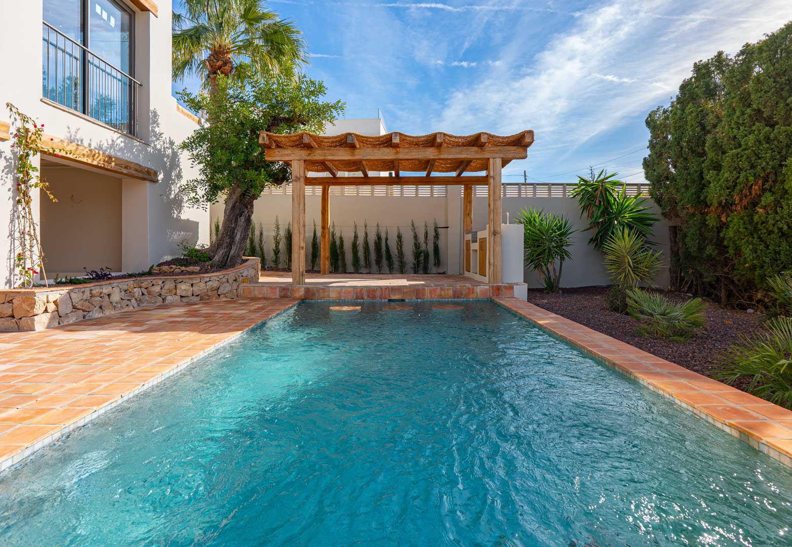 Piscina rectangular con baldosas de terracota, pérgola de madera y jardín mediterráneo con muros de piedra natural y vegetación autóctona bajo un cielo despejado.