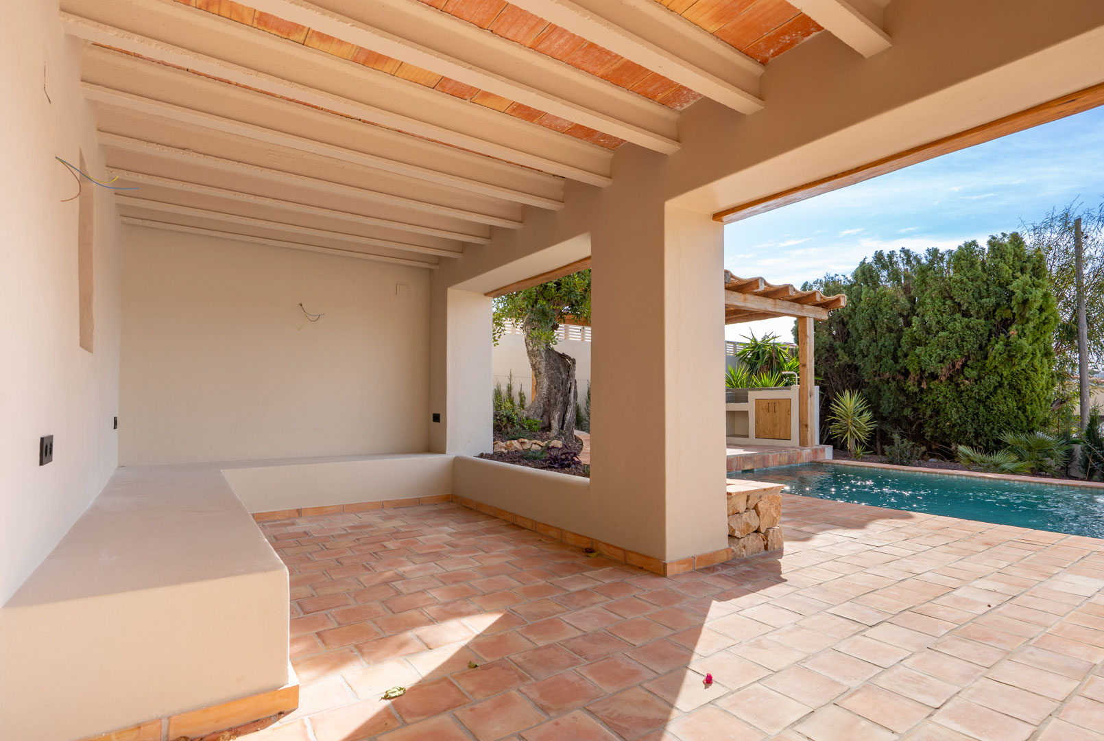 Terraza cubierta con suelos de terracota, techo de vigas vistas y banco de obra, situada junto a la piscina y la zona de pérgola exterior.