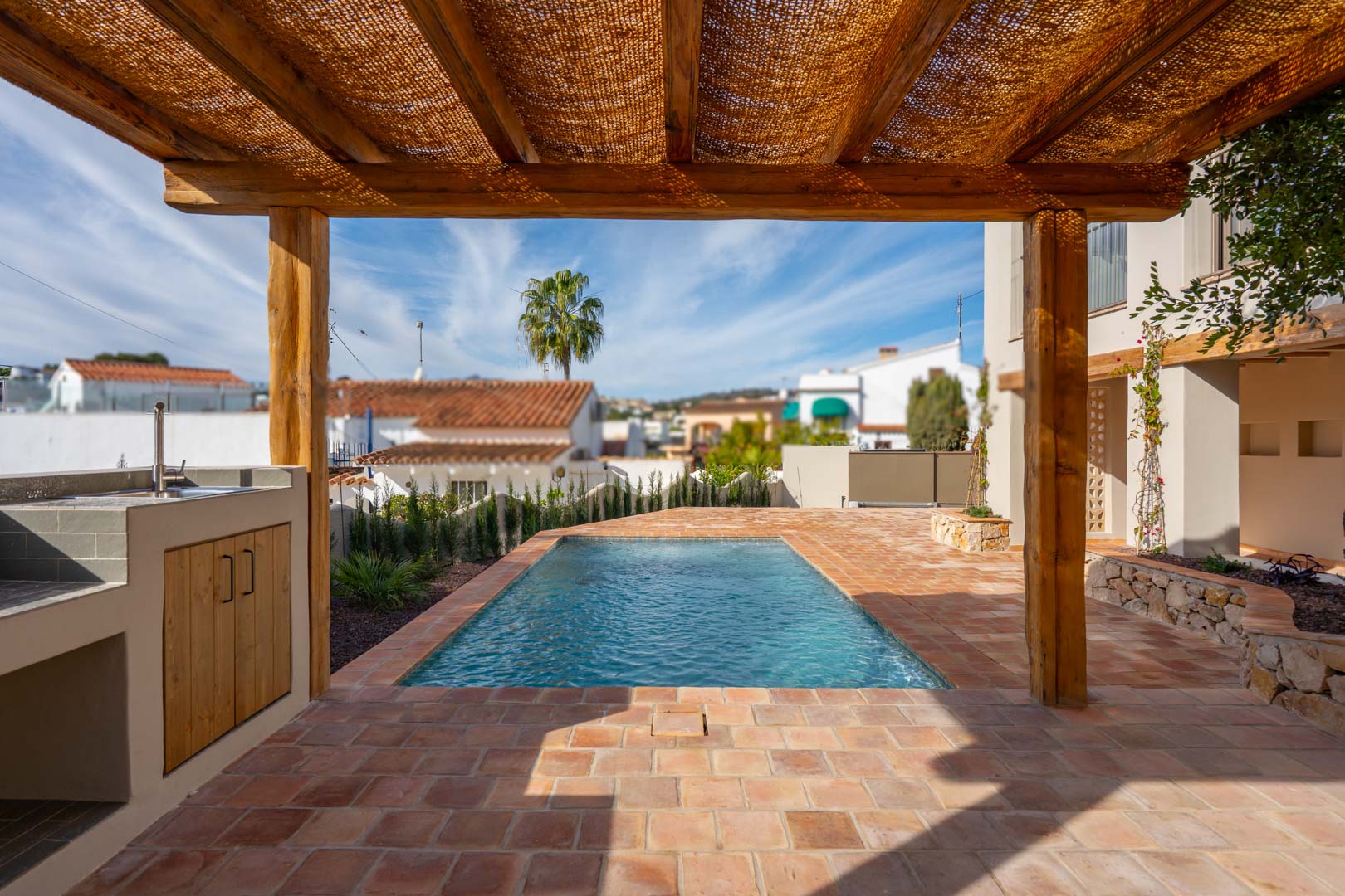 Terraza exterior con suelos de terracota, pérgola de madera y zona de barbacoa junto a la piscina de nueva construcción en un entorno mediterráneo.