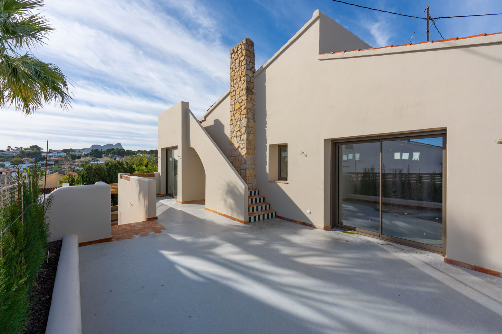 Terraza superior con suelo de hormigón pulido, chimenea de piedra natural y vistas lejanas al Peñón de Ifach bajo un cielo despejado.