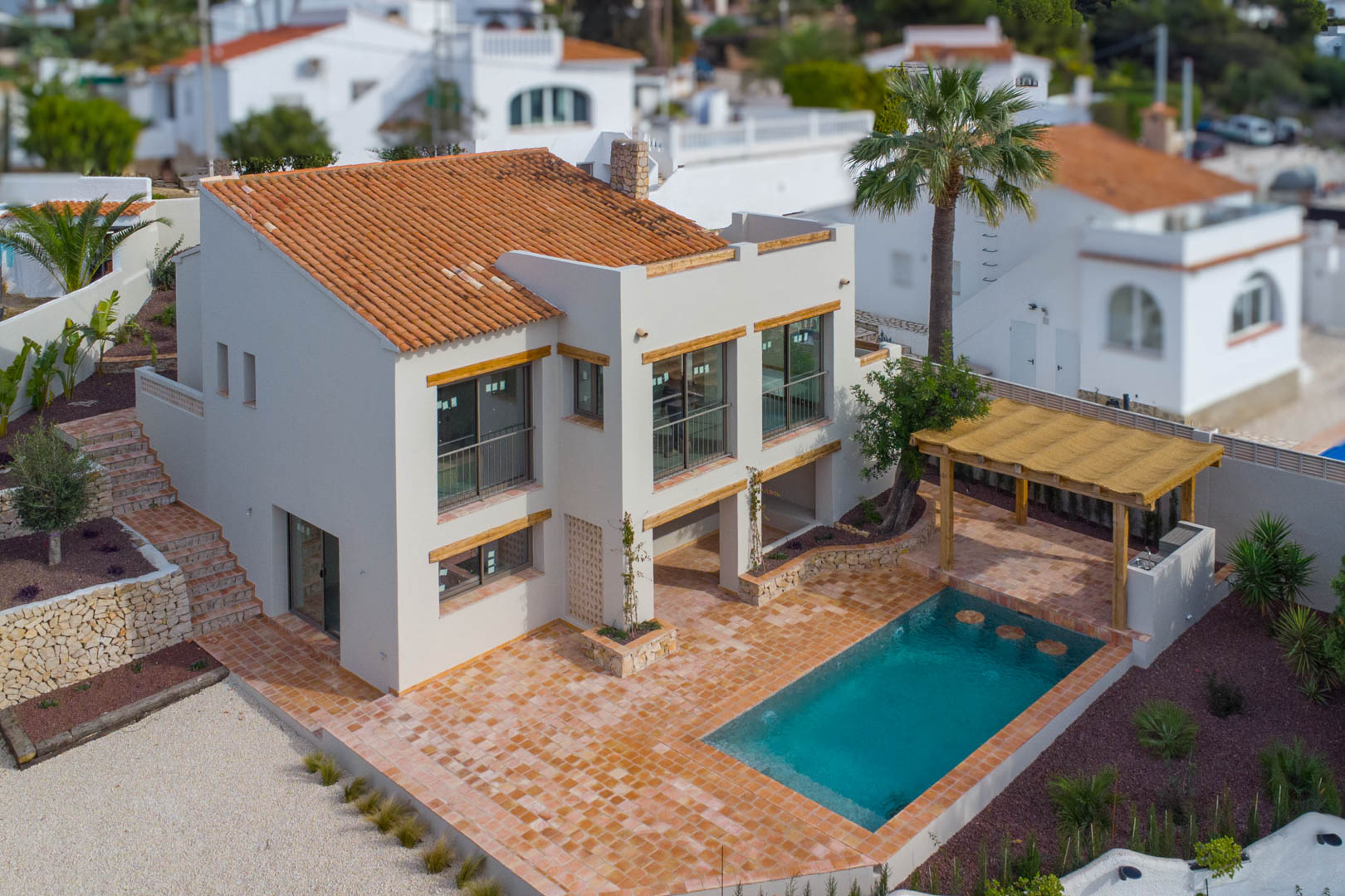 Vista aérea de la villa con piscina de nueva construcción, amplia terraza de terracota y pérgola de madera con zona de barbacoa integrada en el jardín.
