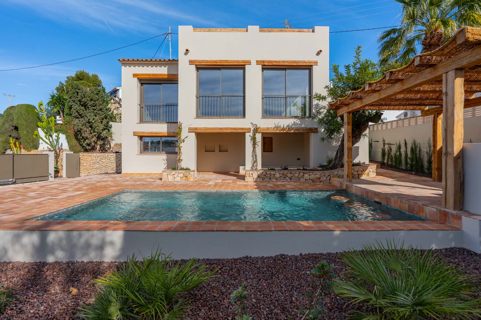 Vista exterior de la villa con piscina rectangular, terraza de baldosas de terracota y una pérgola de madera con cubierta natural junto al jardín mediterráneo.