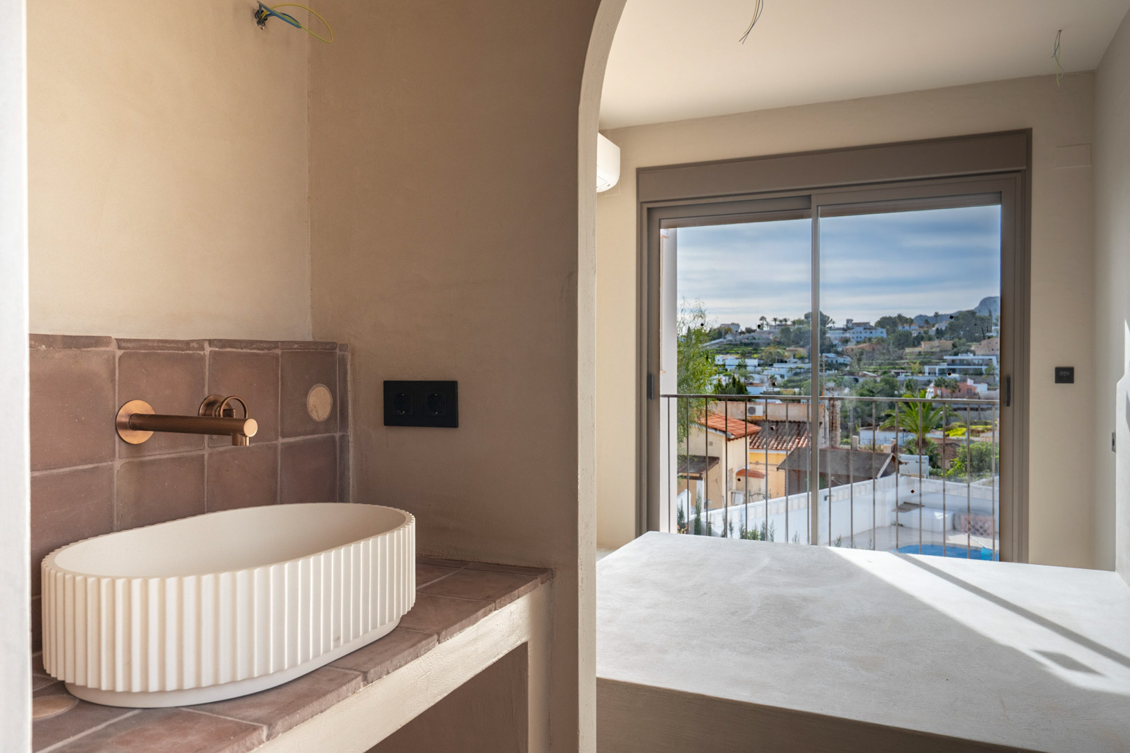 Zona de baño con lavabo sobre encimera estriado y grifería en cobre, conectada mediante un arco al dormitorio con acceso al balcón y vistas despejadas.