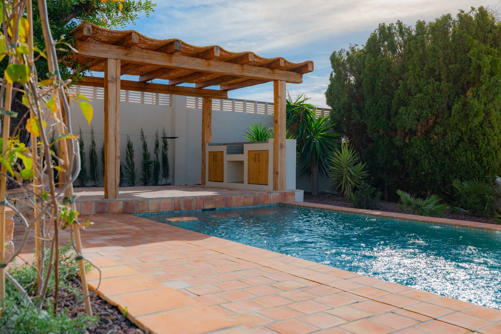 Zona exterior con piscina de mosaico azul, rodeada de suelos de terracota y una pérgola de madera con cocina exterior integrada y vegetación mediterránea.