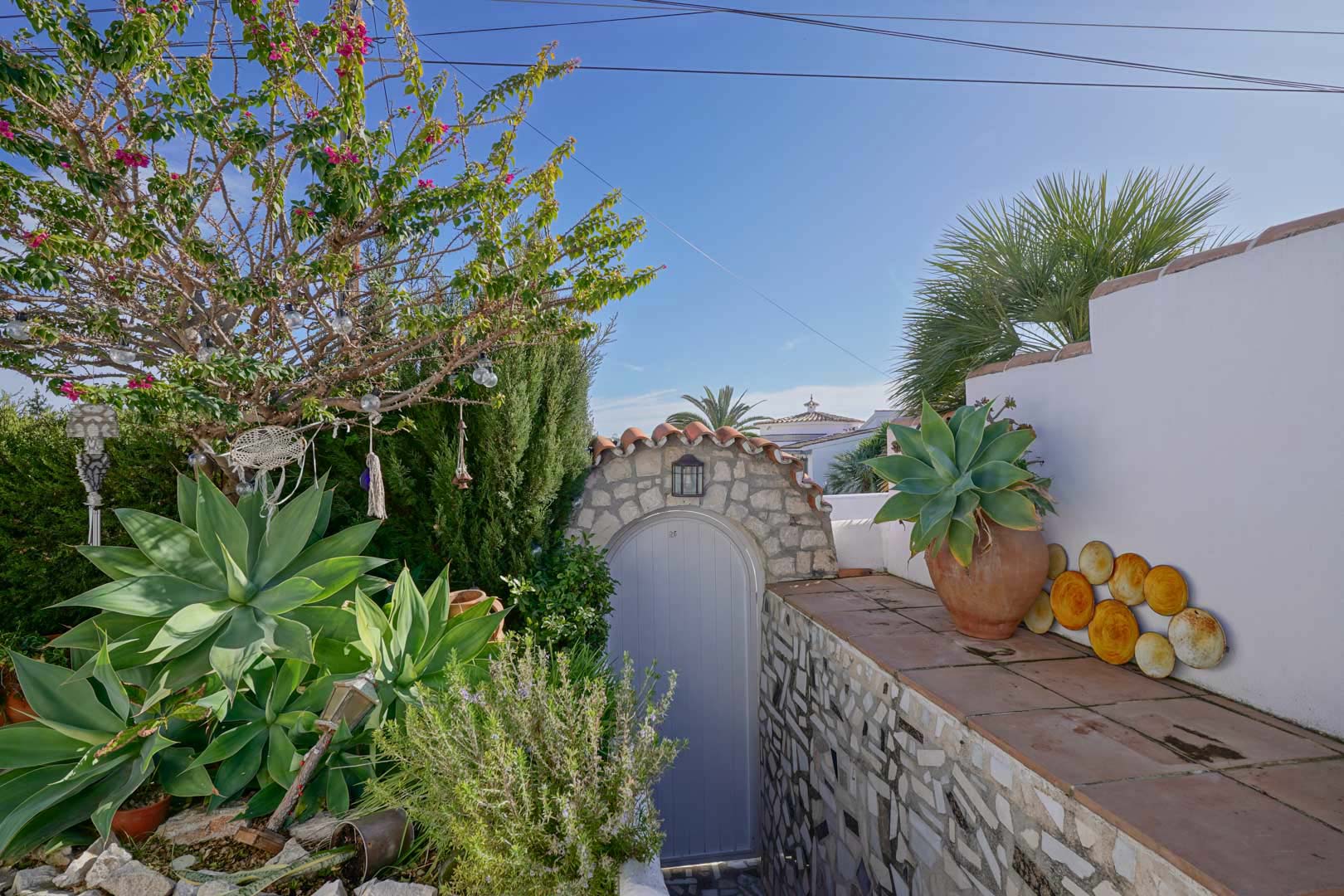 Entrada exterior con arco de piedra natural, puerta en tono claro y vegetación mediterránea autóctona sobre muros de mampostería y baldosas de barro.