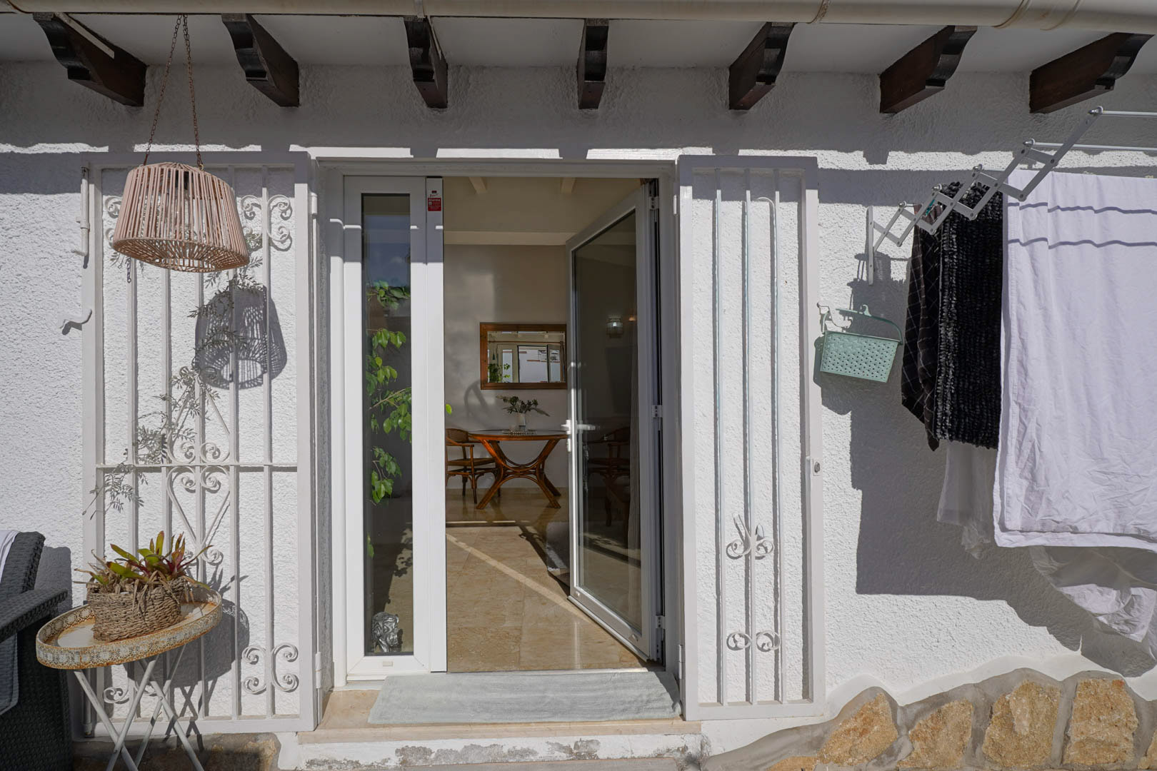 Entrada exterior con puerta de cristal blanca y rejas decorativas bajo vigas de madera. Se aprecia el salón-comedor interior y detalles funcionales en la fachada de estilo mediterráneo.