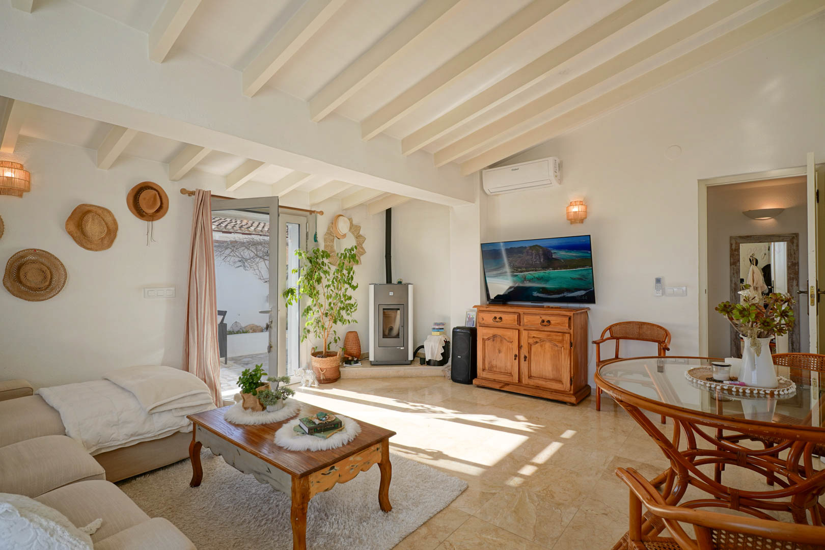 Salón-comedor con techos de vigas de madera blancas, estufa de pellets y suelos de mármol. El espacio cuenta con aire acondicionado y acceso directo a la terraza exterior.