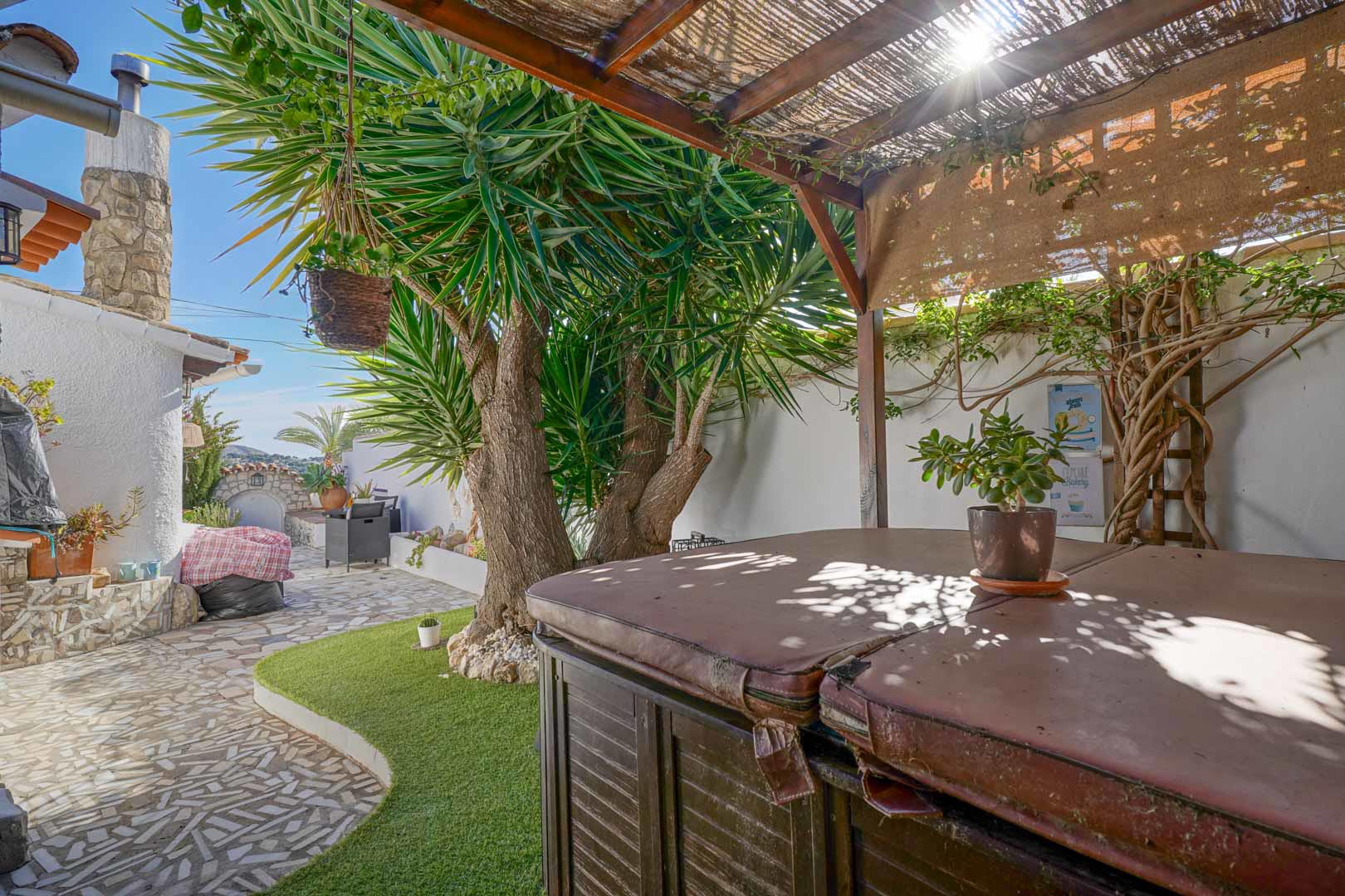 Terraza exterior con jacuzzi cubierto bajo una pérgola de madera, integrada en un jardín de bajo mantenimiento con suelos de piedra y vegetación mediterránea.
