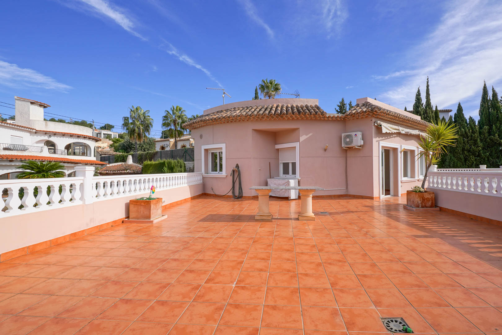 Amplia terraza superior con suelo de baldosas de terracota, balaustrada blanca y mesa de piedra, rodeada de vistas despejadas y arquitectura mediterránea.