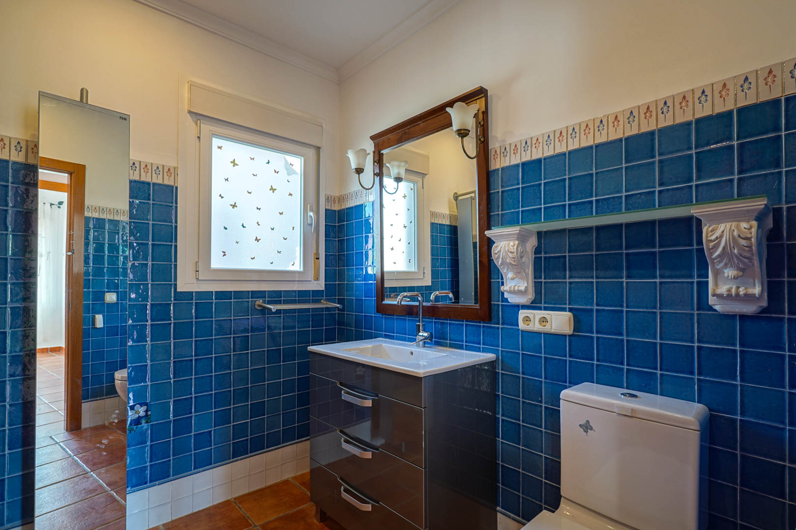 Baño con azulejos azules y cenefa decorativa, mueble de lavabo moderno en tono oscuro, espejo con marco de madera y suelos de terracota tradicionales.