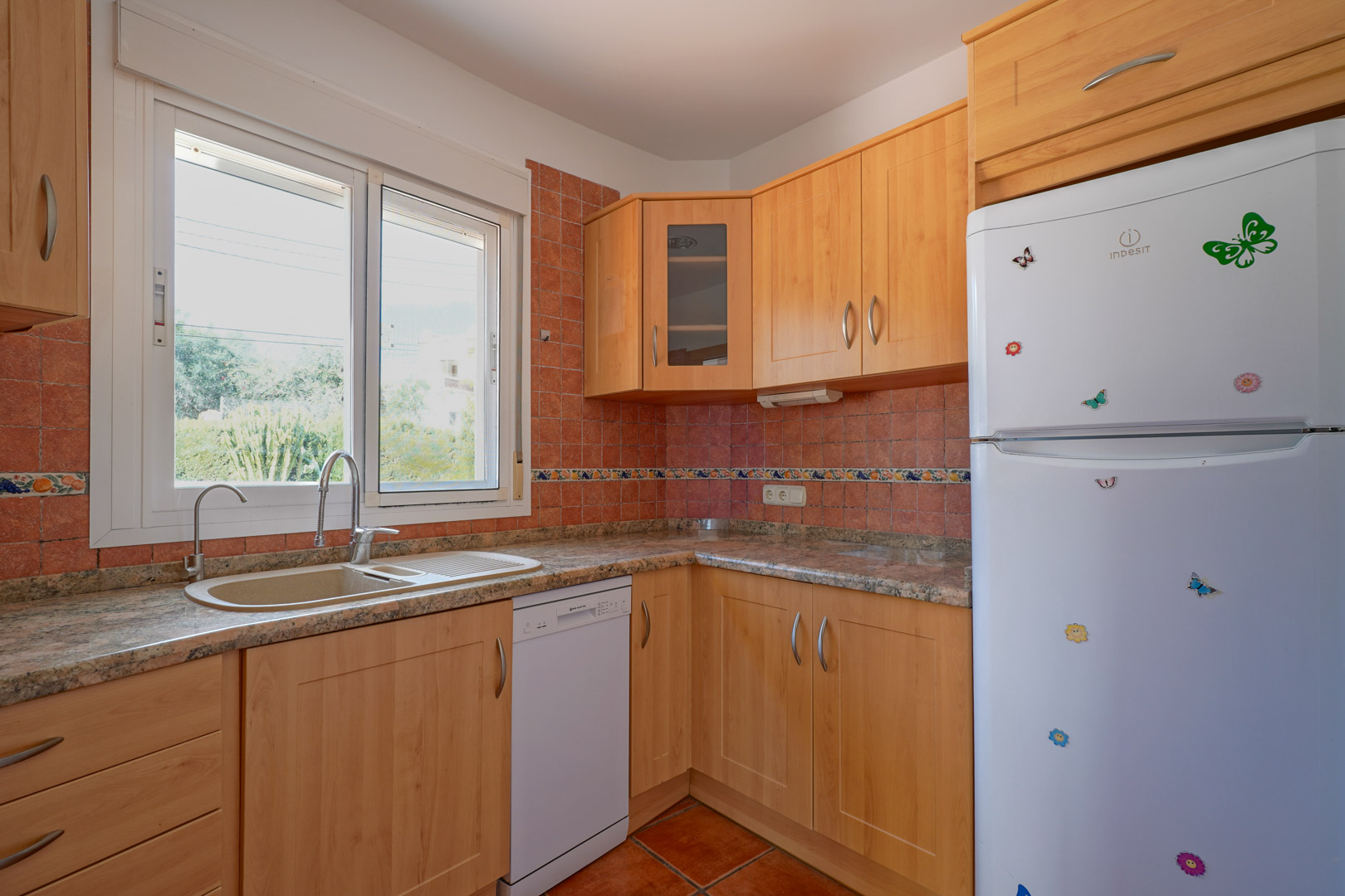 Cocina independiente con mobiliario de madera clara, encimera de granito y azulejos rústicos. Incluye lavavajillas, frigorífico blanco y ventana al exterior con luz natural.