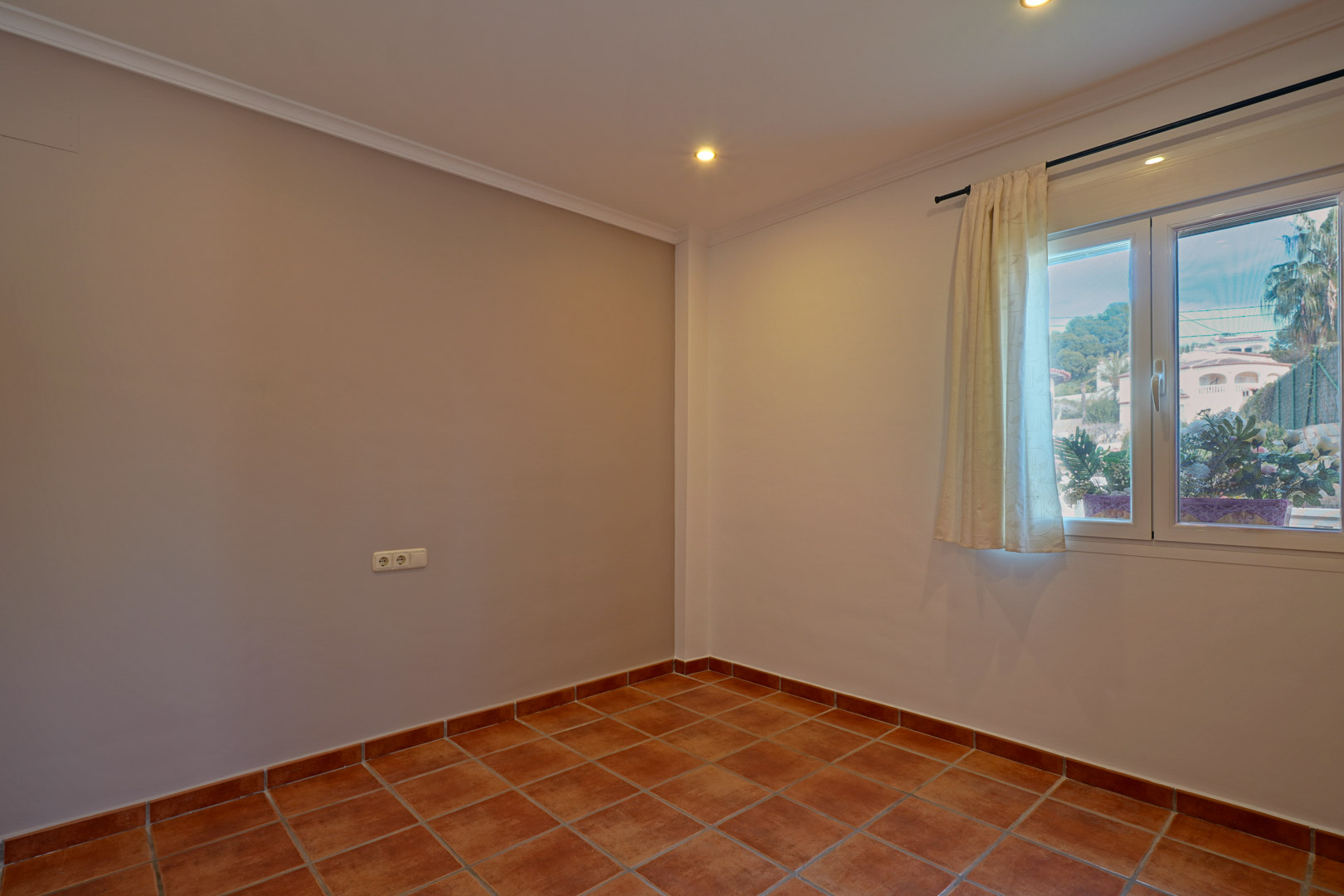 Dormitorio luminoso con suelos de gres rústico, paredes en tonos neutros y ventana de PVC con vistas al entorno natural de Benissa. Incluye iluminación empotrada.