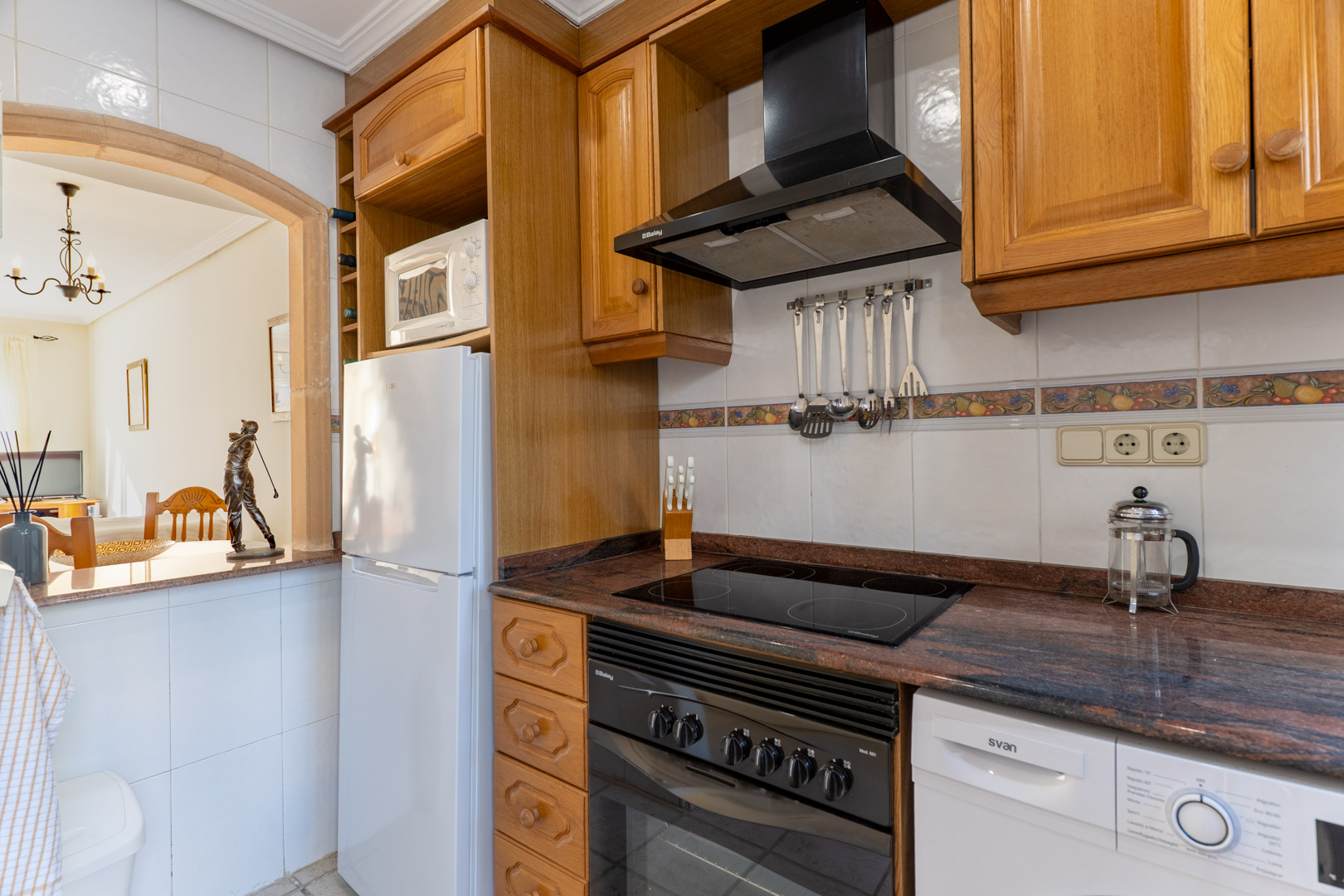 Cocina funcional con armarios de madera, encimera de granito y electrodomésticos, comunicada con el salón a través de un arco decorativo.
