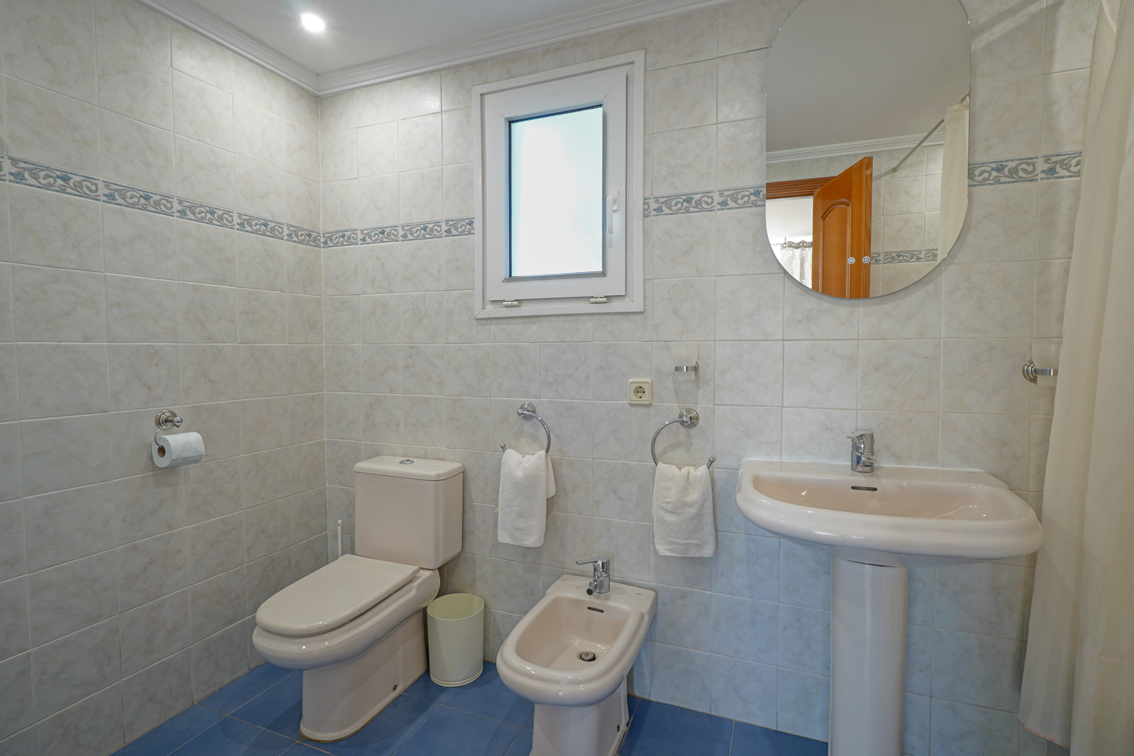 Cuarto de baño completo con azulejos claros, cenefa decorativa y suelo azul. Incluye sanitario, bidé, lavabo de pedestal y una ventana para ventilación natural.