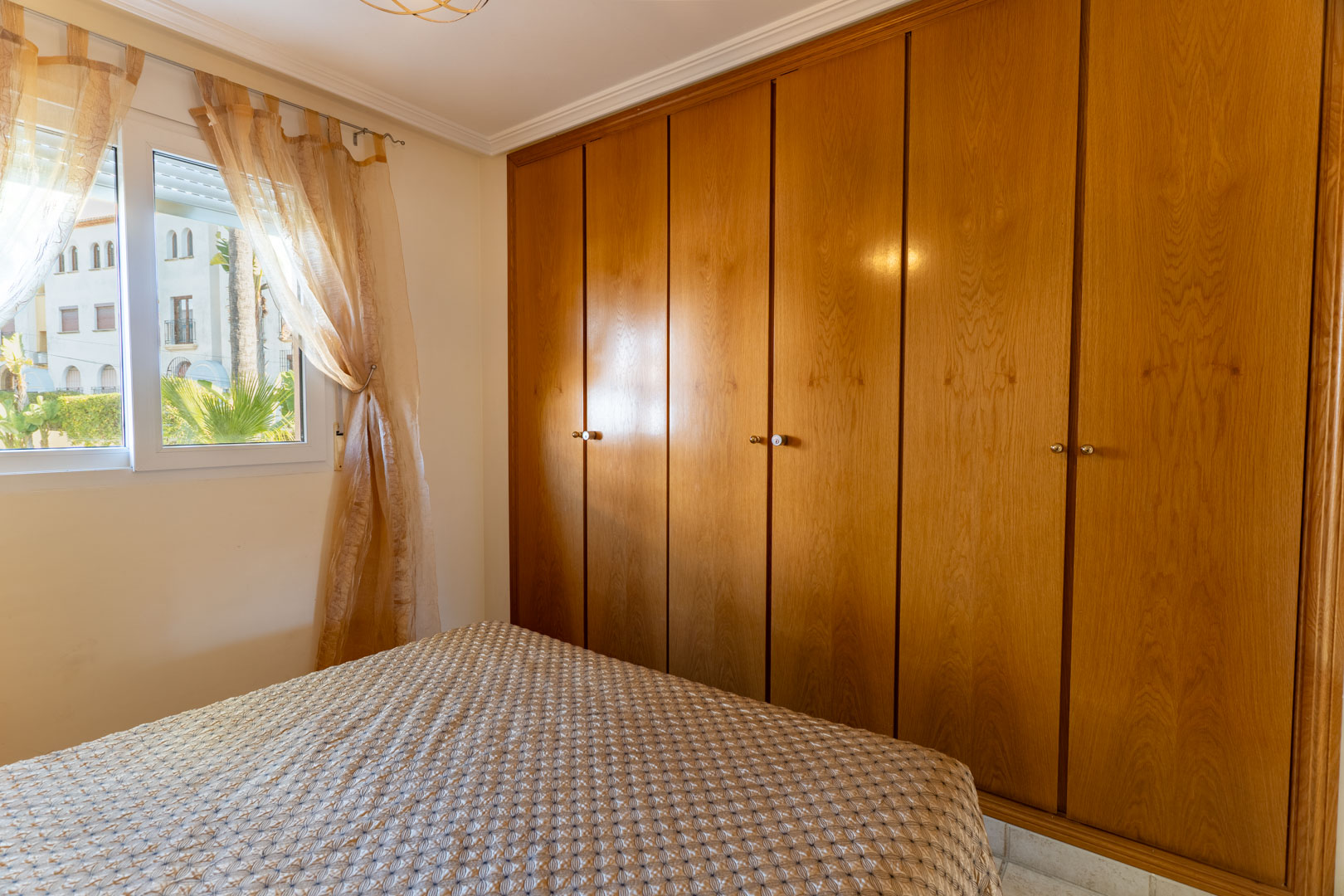Dormitorio con amplio armario empotrado de madera de seis puertas y ventana con vistas al exterior ajardinado, proporcionando luz natural y funcionalidad.