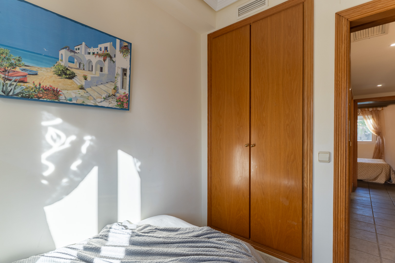 Dormitorio con armario empotrado de madera y luz natural. La estancia conecta a través de un marco de madera con un pasillo distribuidor de suelos cerámicos.