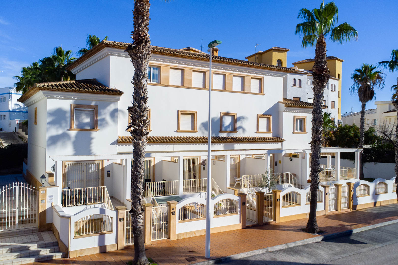 Fachada exterior de viviendas tríplex de estilo mediterráneo con paredes blancas, marcos de piedra y terrazas privadas valladas frente a la calle.