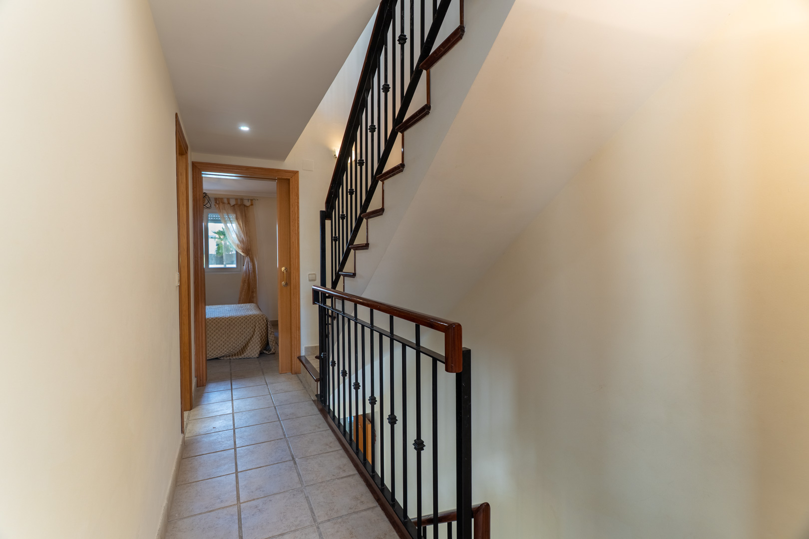 Pasillo distribuidor con suelos cerámicos que conecta con los dormitorios, destacando la escalera con barandilla de madera y forja negra.
