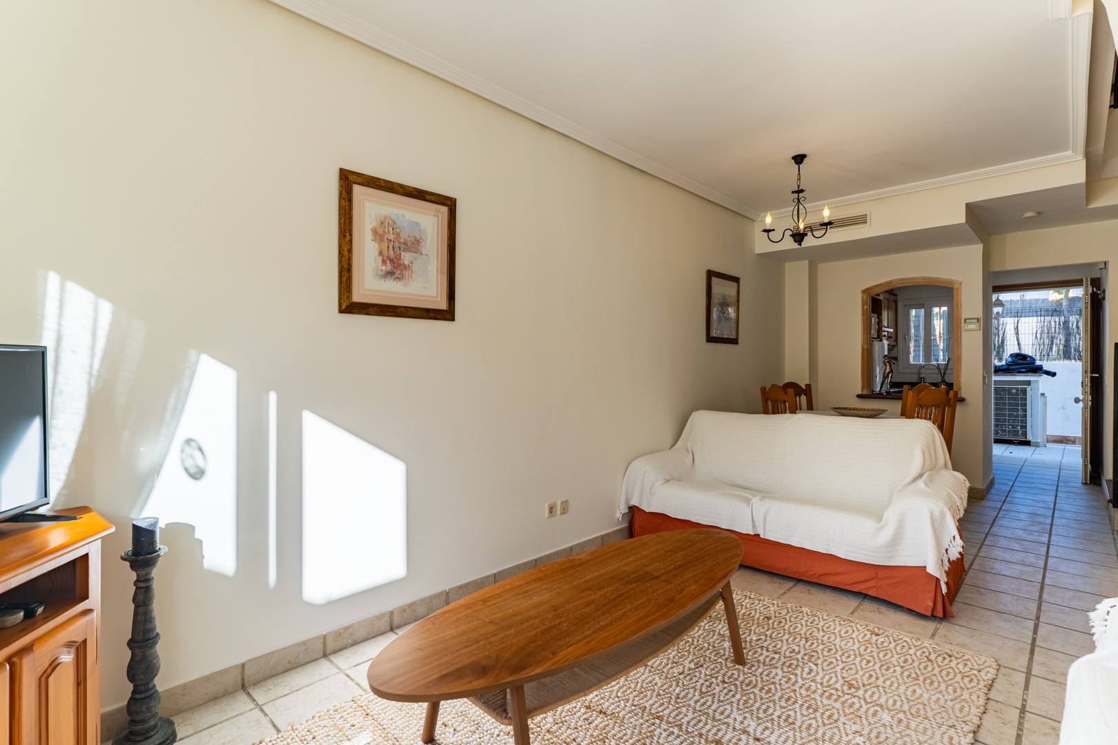 Salón comedor luminoso con suelos cerámicos, mesa de centro de madera y apertura hacia la cocina, conectando visualmente con el patio exterior.