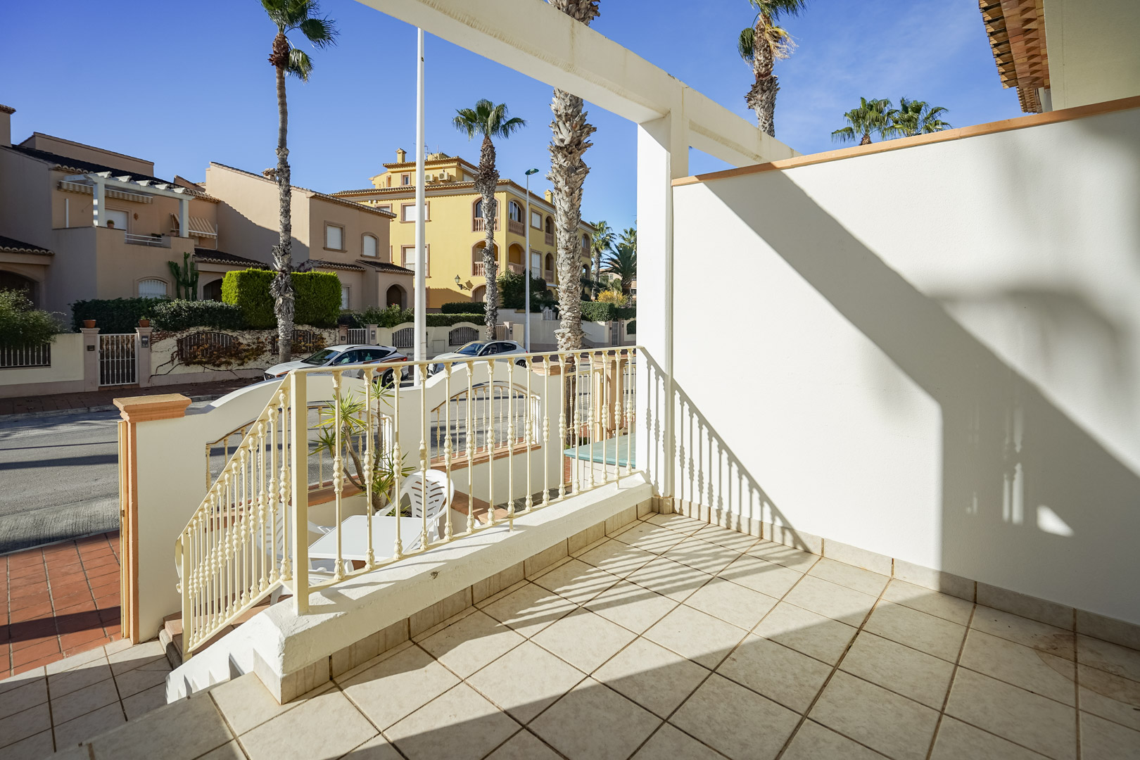 Terraza delantera elevada con suelos cerámicos y barandilla metálica, con vistas a la calle Génova y las palmeras del entorno residencial de Jávea.