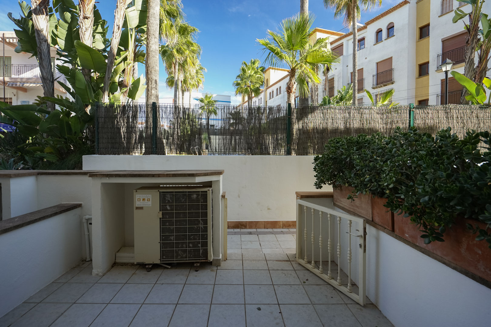 Terraza privada con suelo cerámico, jardinera integrada y recinto para unidad de aire acondicionado, rodeada de vegetación tropical y palmeras.
