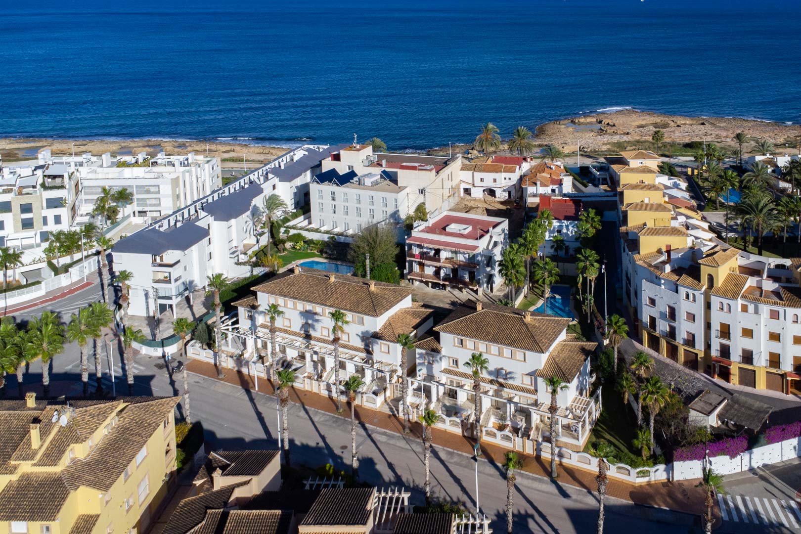 Vista aérea del complejo en Calle Génova, mostrando su cercanía al mar Mediterráneo y la arquitectura de fachadas blancas con tejados de teja rodeadas de palmeras.