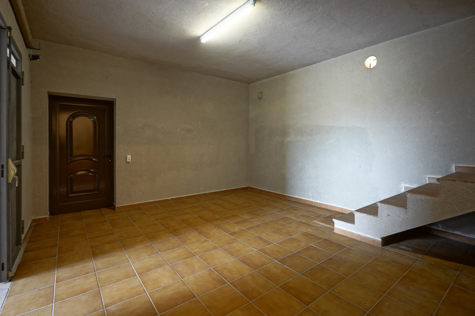 Amplia estancia con suelos de gres rústico, paredes de acabado rugoso, acceso a escalera lateral y puerta de madera oscura.