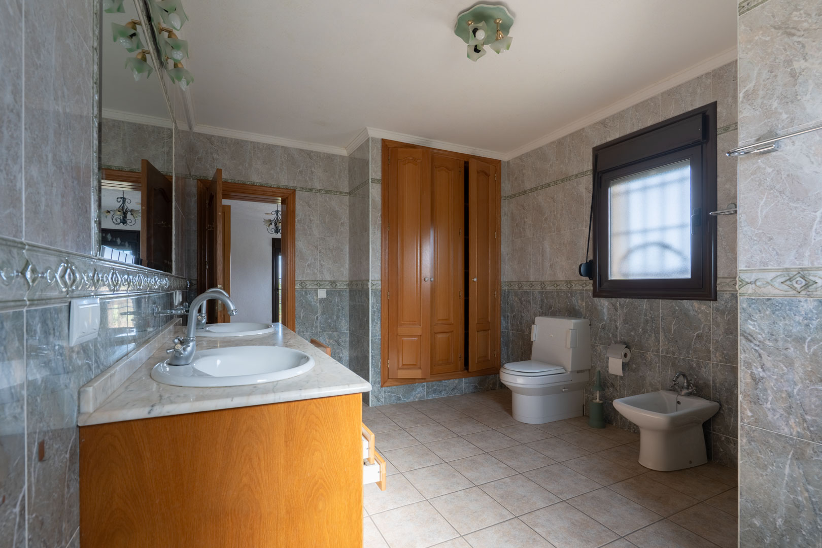 Baño espacioso con lavabo doble de mármol, armarios de madera empotrados y paredes alicatadas con cenefas decorativas. Incluye sanitario y bidé.