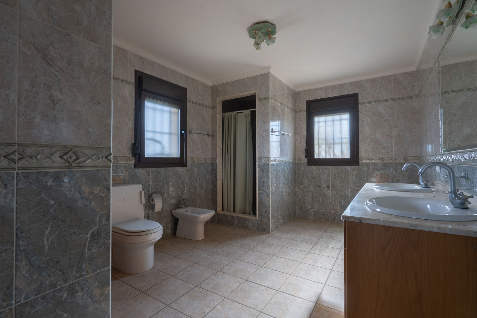 Baño espacioso con mueble de doble lavabo y encimera de mármol, paredes alicatadas en gris con cenefa y dos ventanas con marcos oscuros.