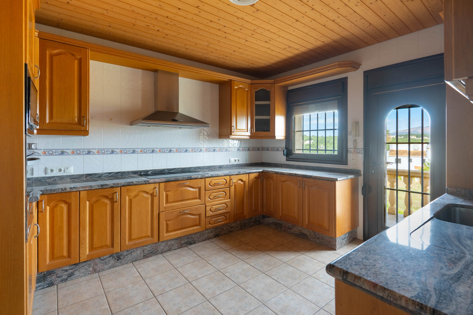 Cocina con armarios de madera, encimeras de granito y techo de madera. Incluye campana de acero inoxidable y puerta con acceso a una terraza con vistas despejadas.
