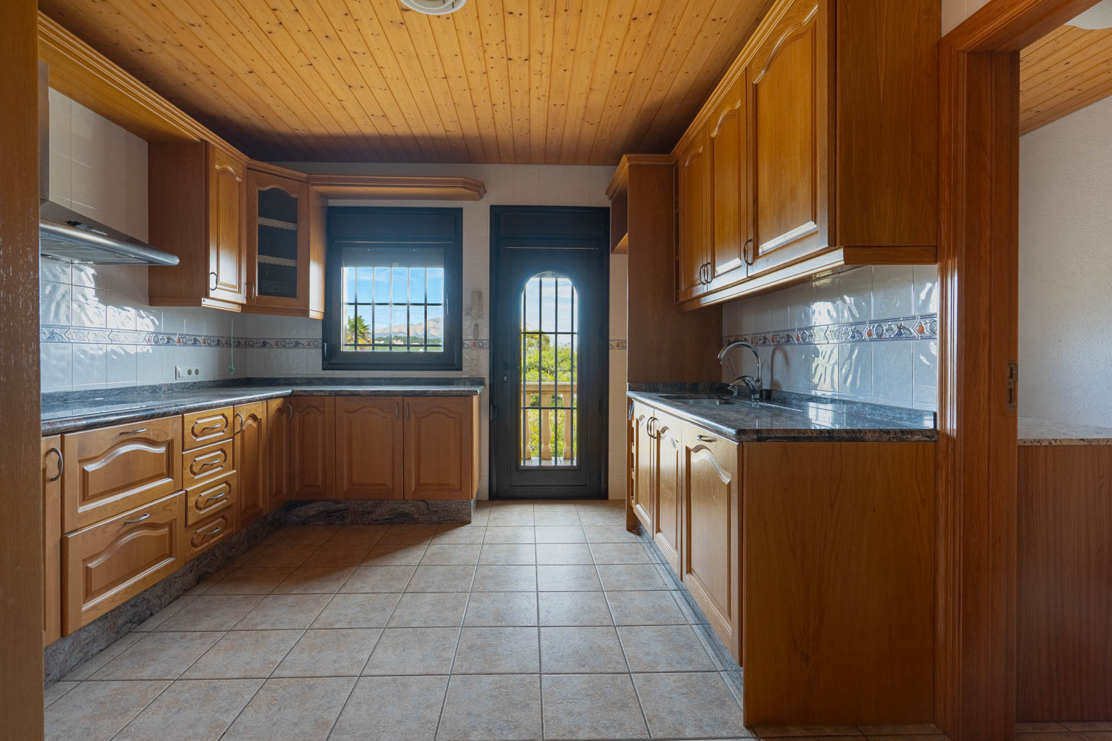 Cocina con mobiliario de madera, encimeras de granito y techo revestido en madera. Incluye suelos cerámicos y acceso directo a un balcón con vistas despejadas.