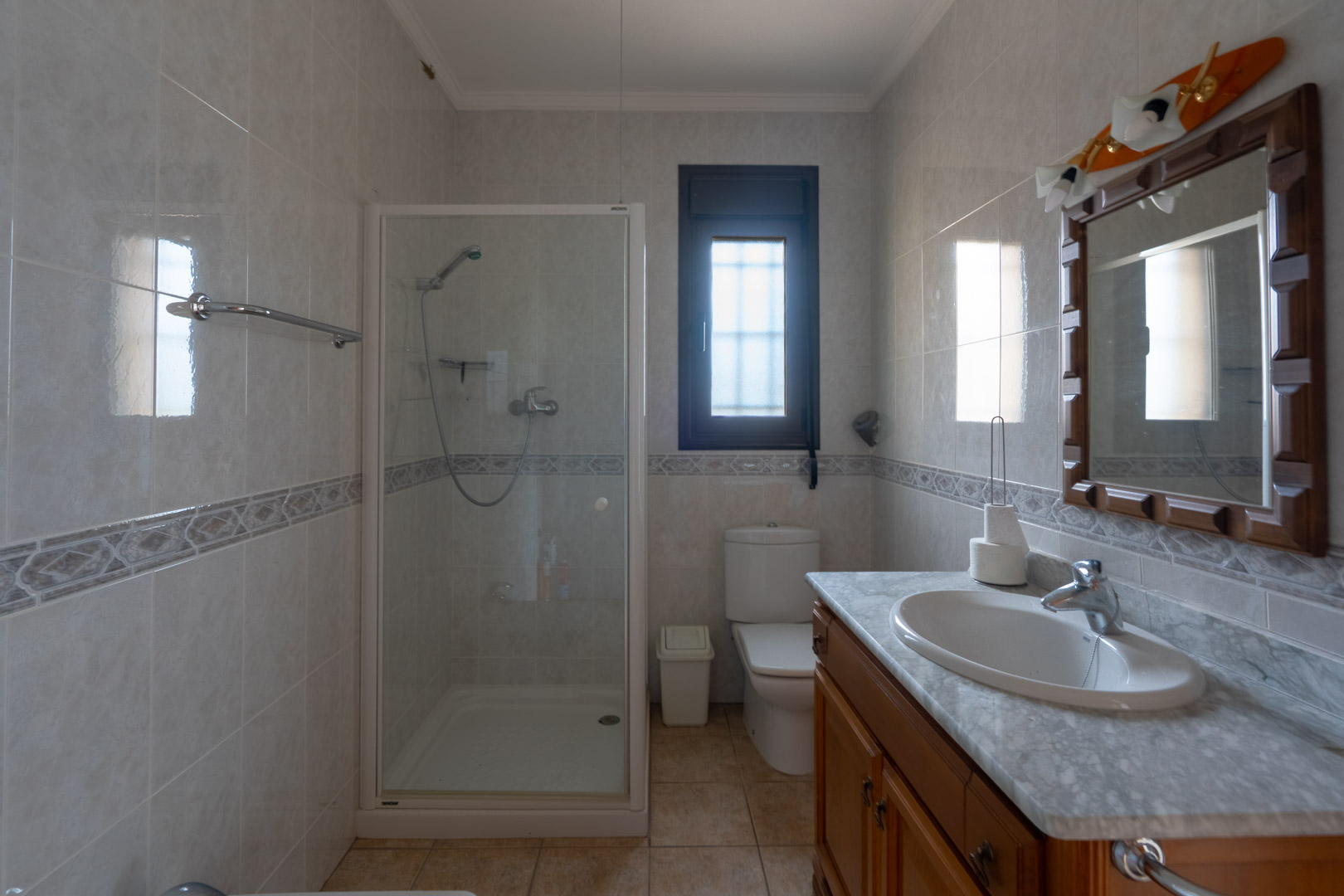 Cuarto de baño con encimera de mármol, mueble de madera y cabina de ducha acristalada. Paredes alicatadas con cenefa decorativa y ventana de cristal translúcido.