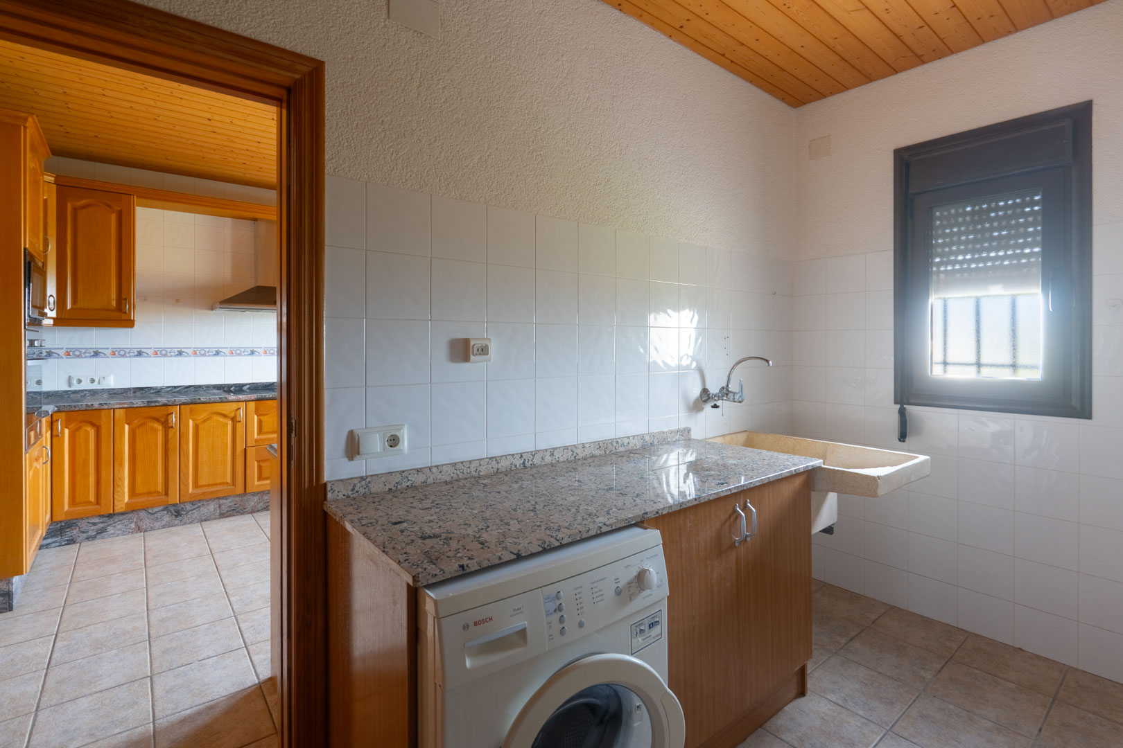 Cuarto de lavado con encimera de granito, lavadora Bosch y fregadero de piedra. Dispone de techos de madera y paredes alicatadas, situado junto a la cocina principal.