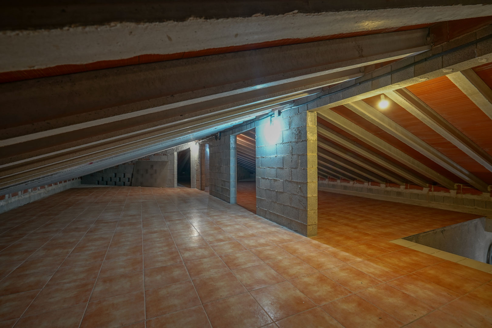 Espacio bajo cubierta con suelo de baldosas cerámicas, vigas de hormigón visto y muros divisorios de bloques de cemento con iluminación funcional.